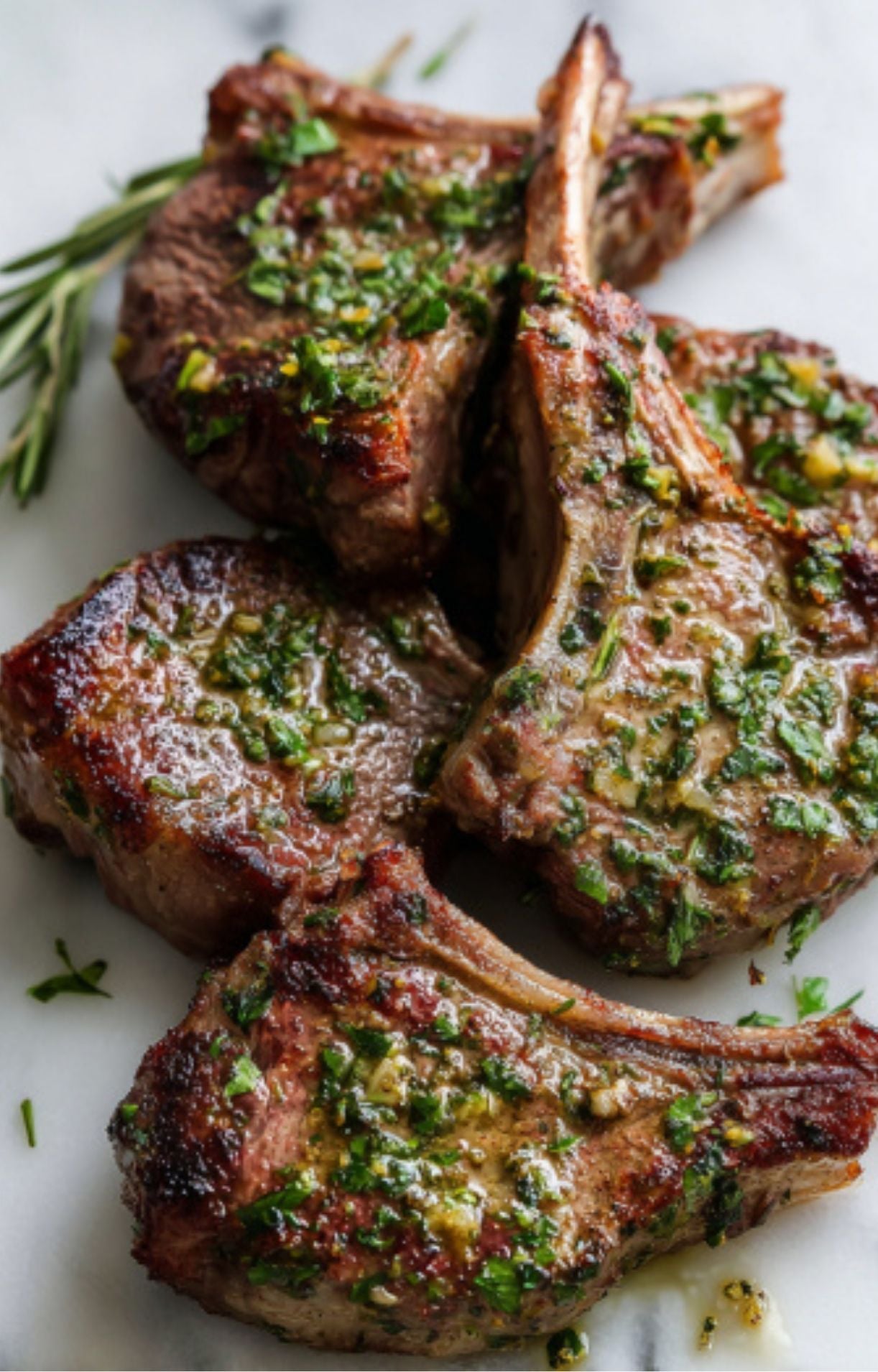 A close-up, angled shot of a stack of juicy lamb chops cooked in the air fryer, heavily topped with a fresh garlic and parsley gremolata on a white marble background.