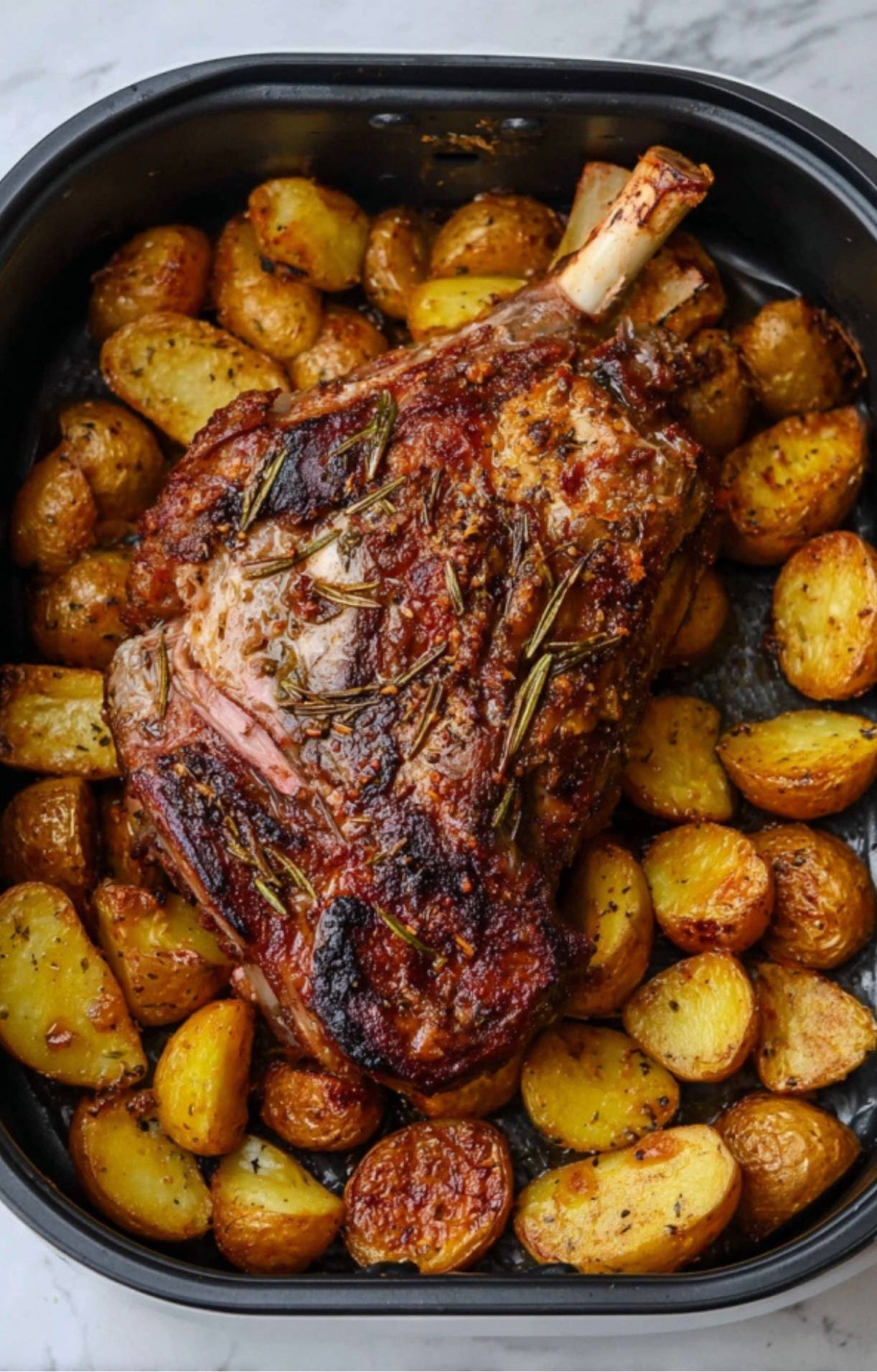 A roasted leg of lamb with a deep herb crust and golden-brown halved potatoes inside a black air fryer basket.