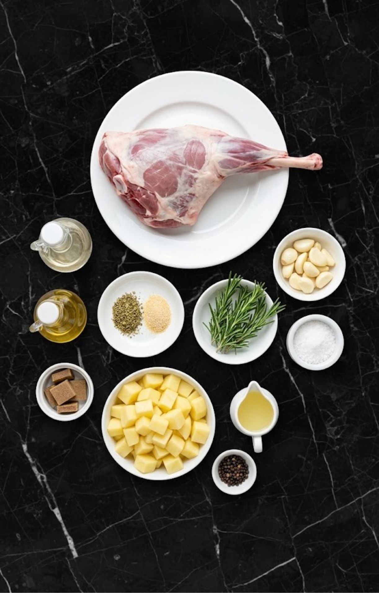 Raw leg of lamb on a white plate surrounded by bowls of diced potatoes, garlic cloves, fresh rosemary, olive oil, and spices on a black marble surface.