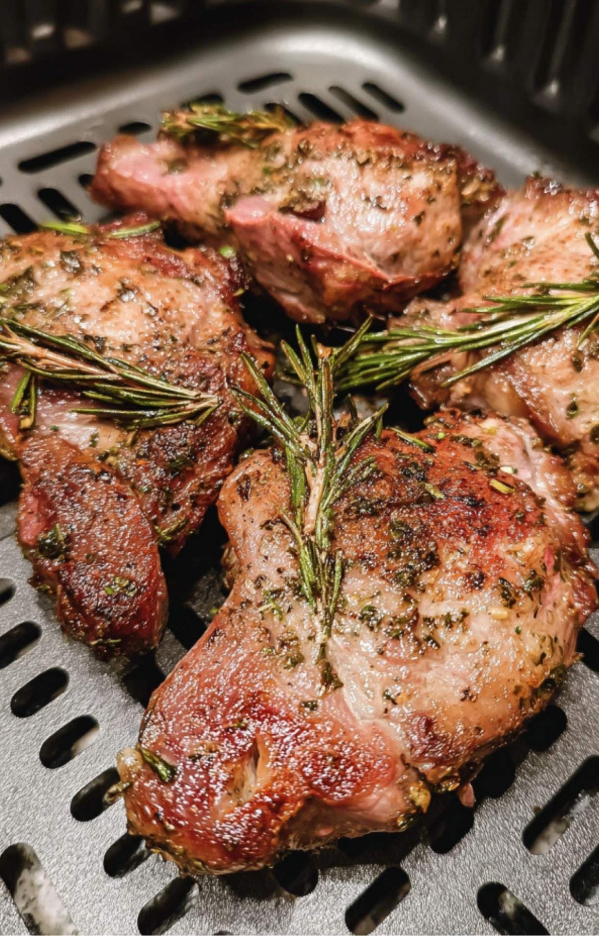 Juicy, browned lamb steaks topped with toasted rosemary sprigs sitting in an air fryer basket after cooking.