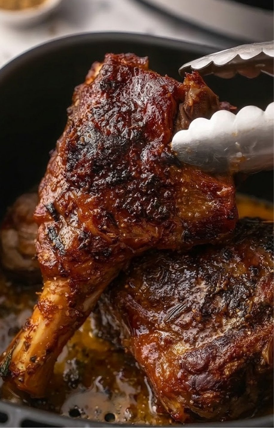 A pair of silver kitchen tongs lifting a tender, caramelized lamb shank from an air fryer basket, revealing a deep golden-brown crust and glistening juices.