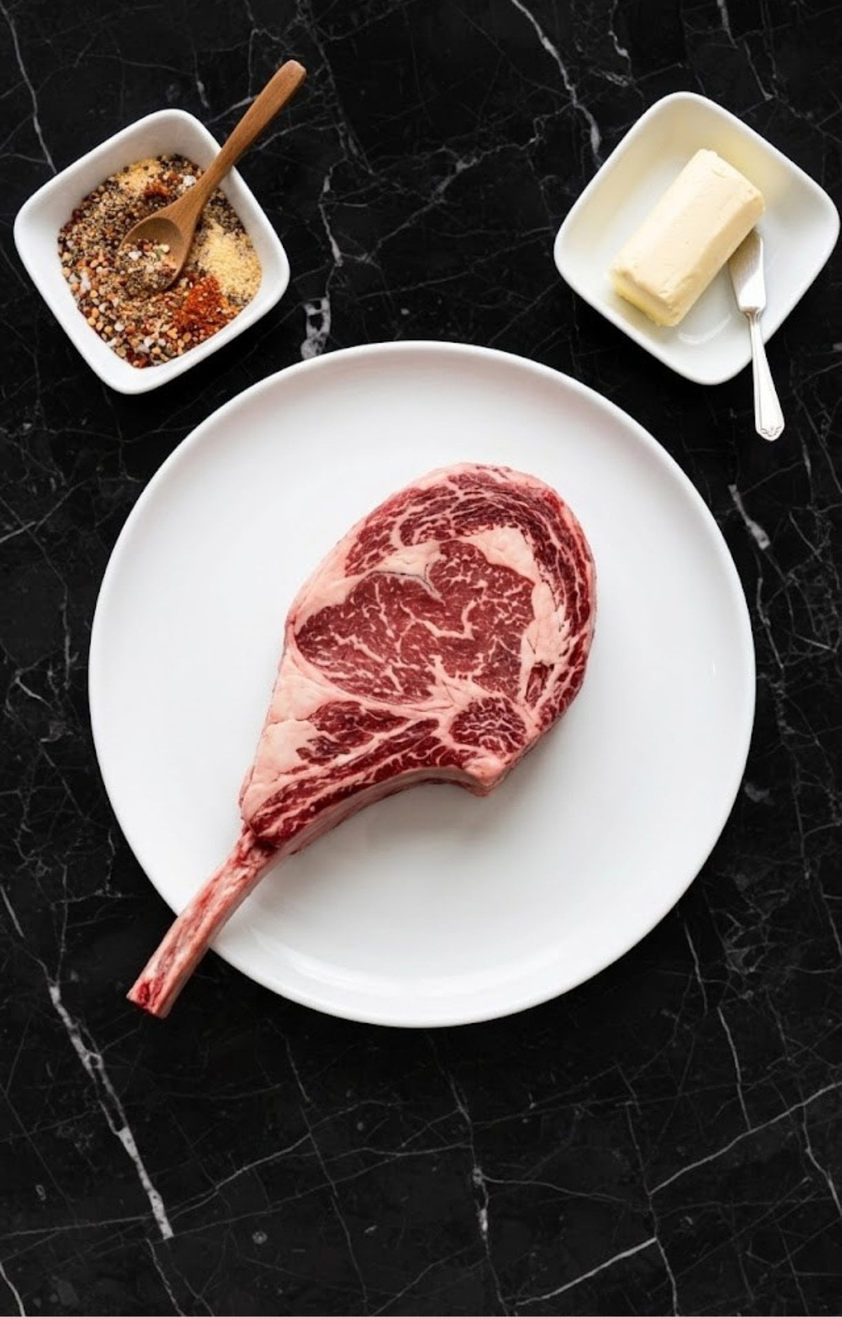 A raw, marbled tomahawk steak on a white plate with bowls of dry rub seasoning and butter.