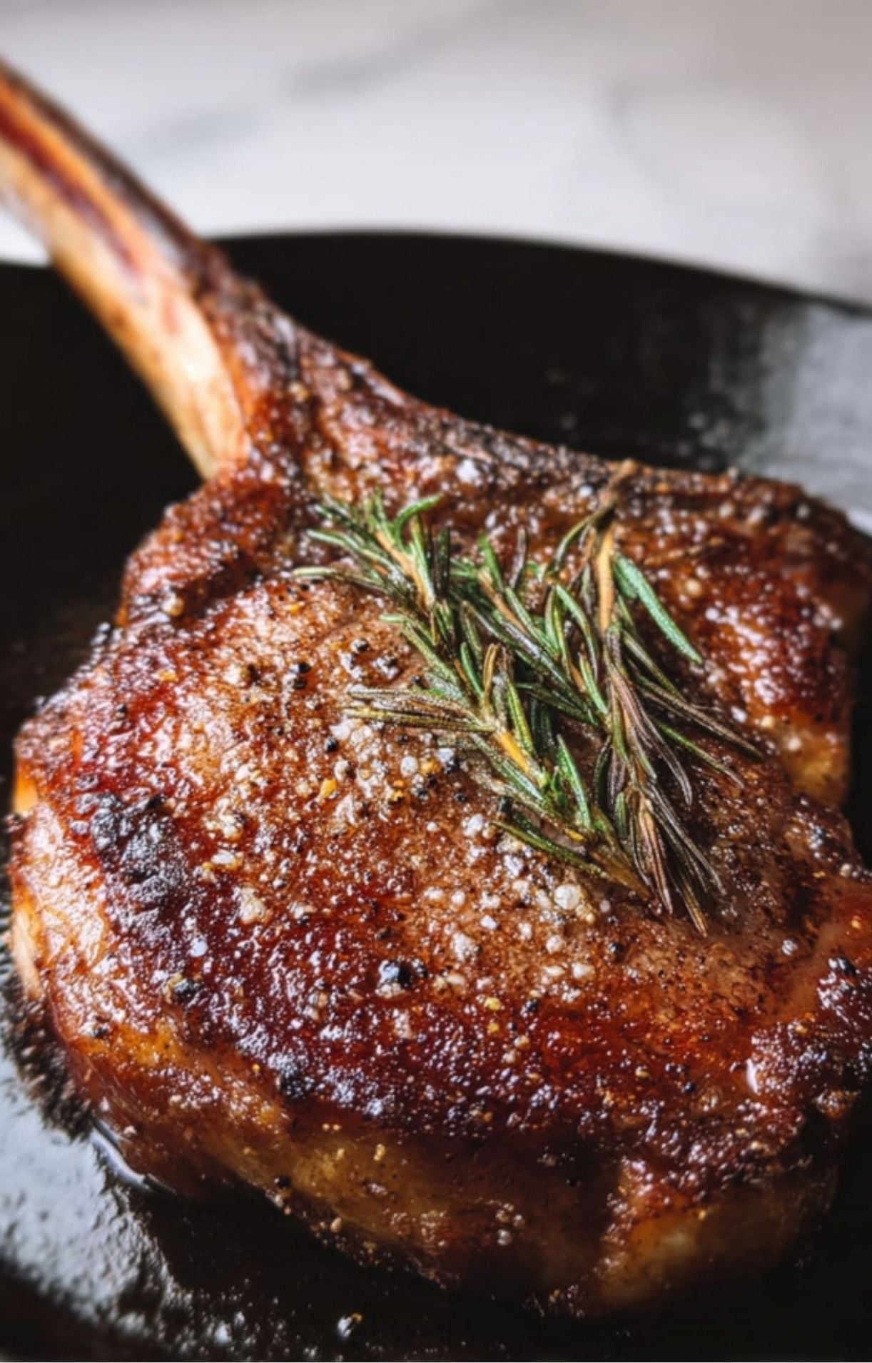 A thick, bone-in tomahawk steak with a deep brown seared crust, topped with a sprig of fresh rosemary and coarse salt in a dark cast-iron skillet.