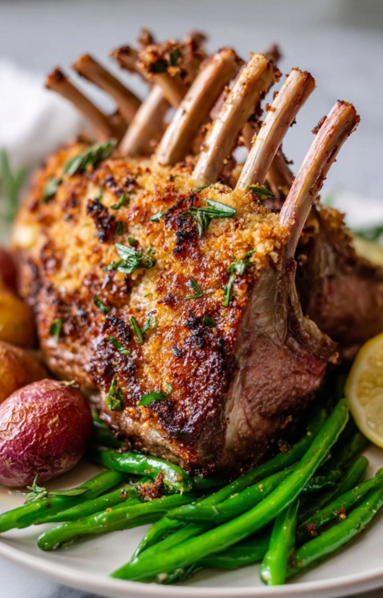 A whole, roasted air fryer rack of lamb with a crispy breadcrumb crust, served on a plate with green beans, red potatoes, and a lemon wedge.