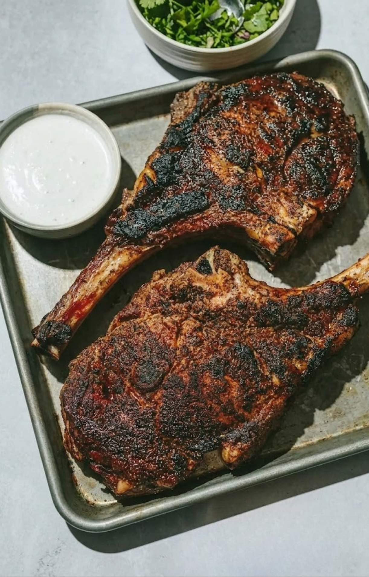 Two whole smoked tomahawk steaks resting on a baking sheet, accompanied by a small bowl of white creamy sauce and a bowl of chopped herbs.