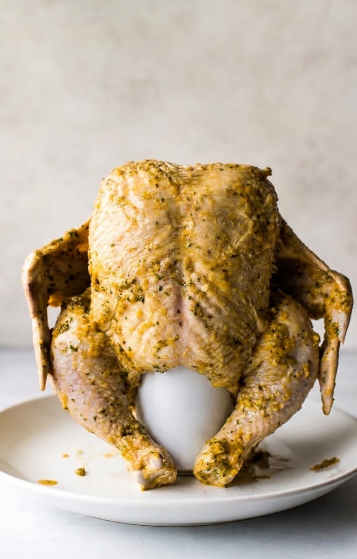 A raw, seasoned chicken propped upright on a white ceramic stand, ready to be roasted for Beer Can Chicken Tacos.
