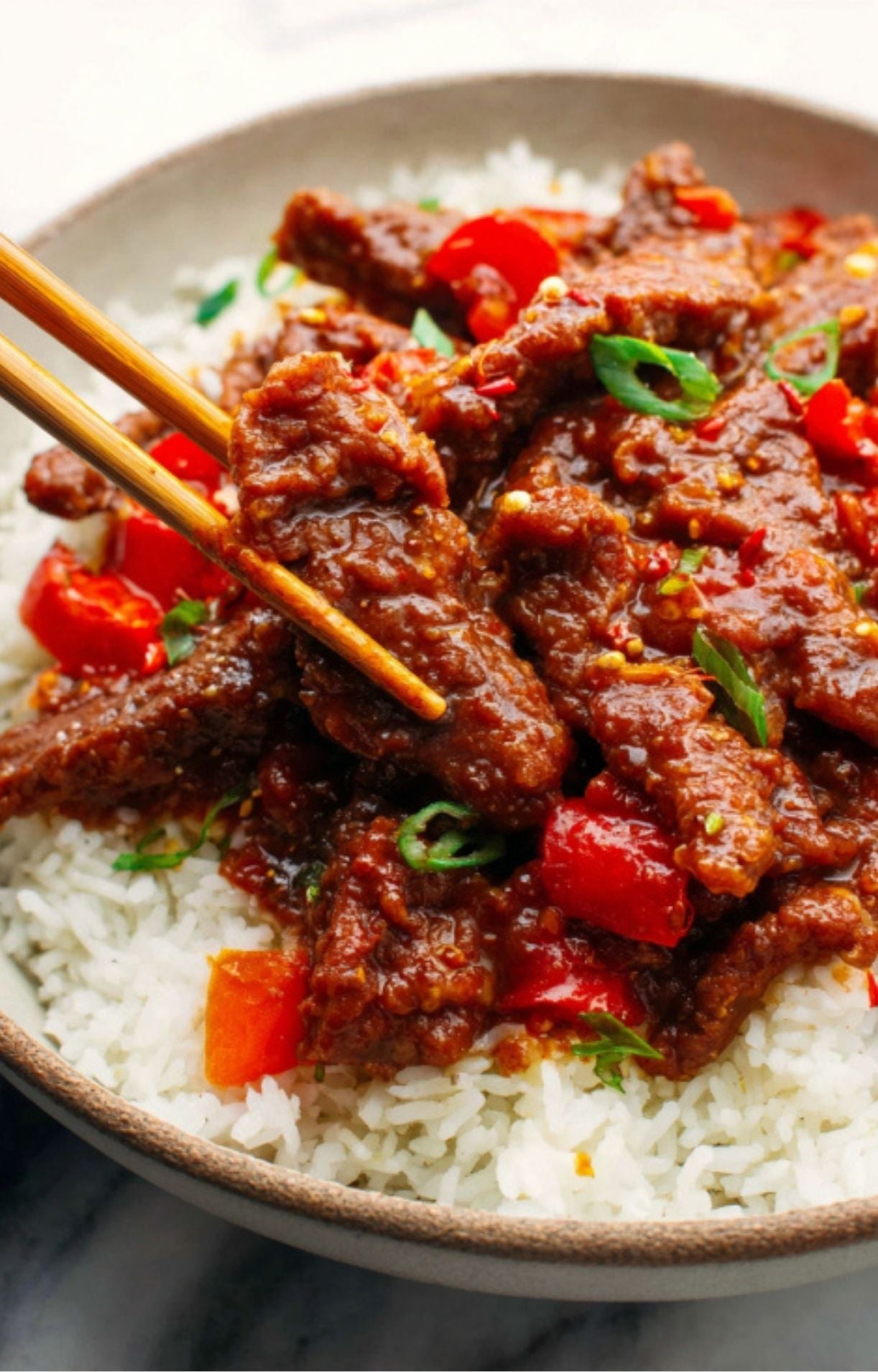 A macro shot of a single piece of saucy beef being lifted by chopsticks from a bowl of white rice.