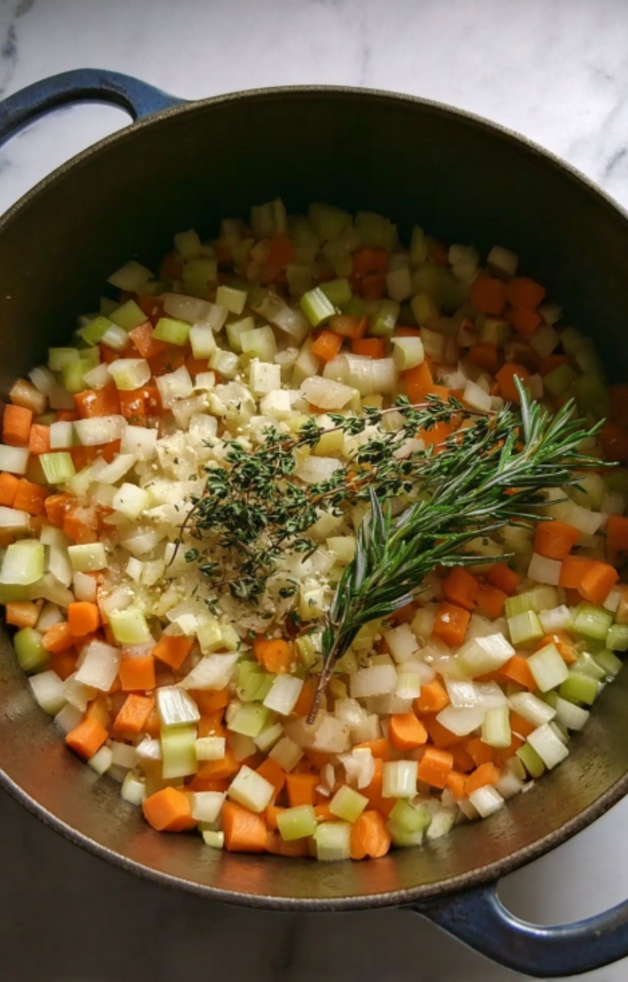 Diced carrots, celery, and onions (mirepoix) sautéing in a pot with fresh rosemary and thyme sprigs to build aromatic flavor.