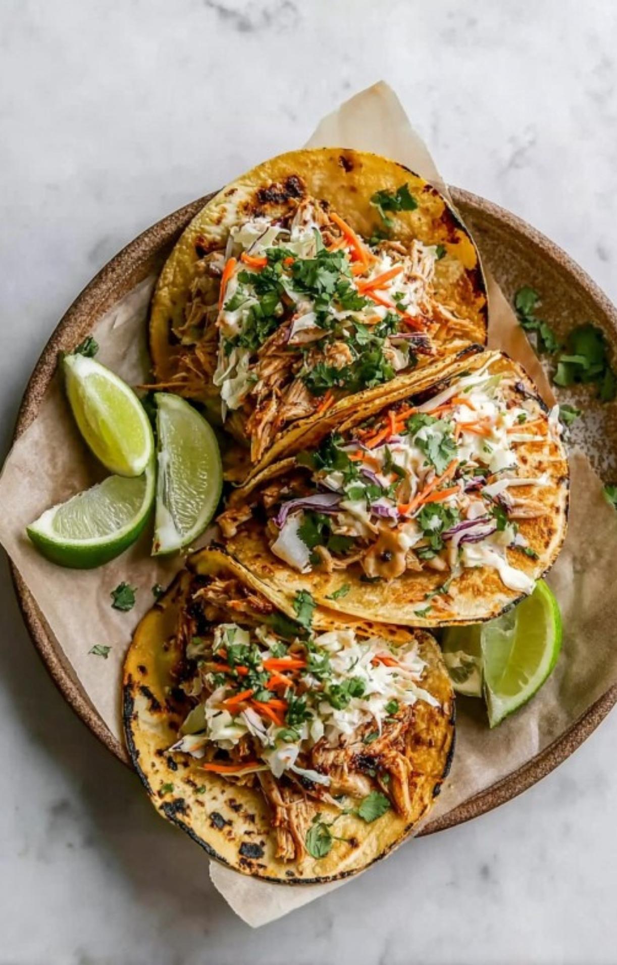 An overhead view of a rustic plate holding three corn tortillas filled with juicy shredded chicken, topped with a colorful cabbage and carrot slaw and fresh cilantro, served with lime wedges.