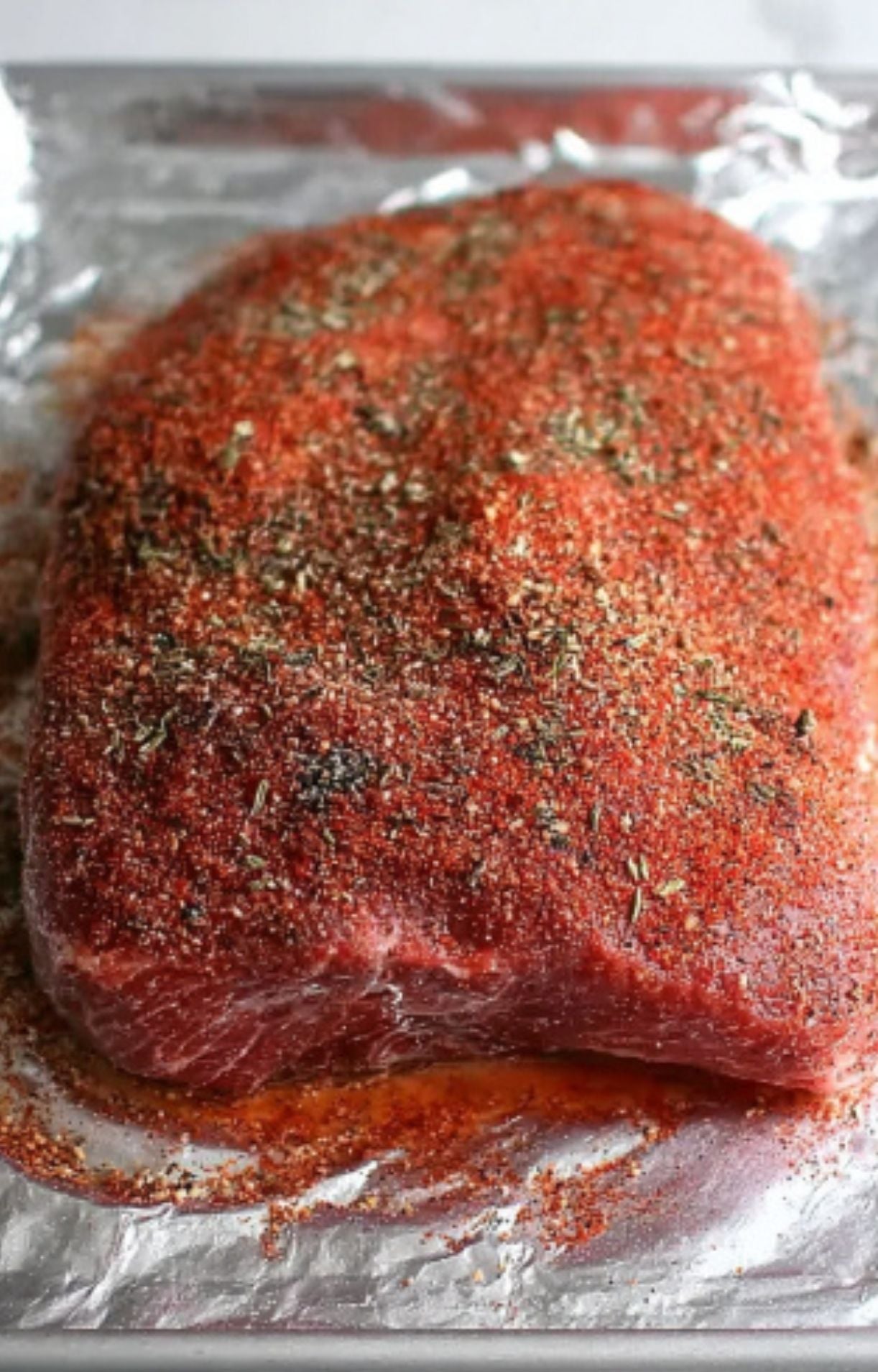 A raw beef brisket placed on aluminum foil and generously coated with a red and herbal dry rub seasoning.
