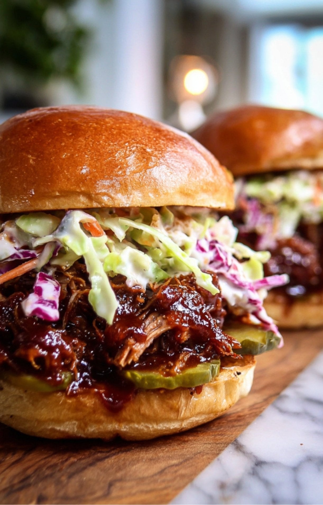 A detailed, eye-level close-up of a smoked brisket sandwich featuring shredded meat tossed in barbecue sauce, topped with creamy coleslaw and pickles, served on a toasted brioche bun over a white marble and wood surface.