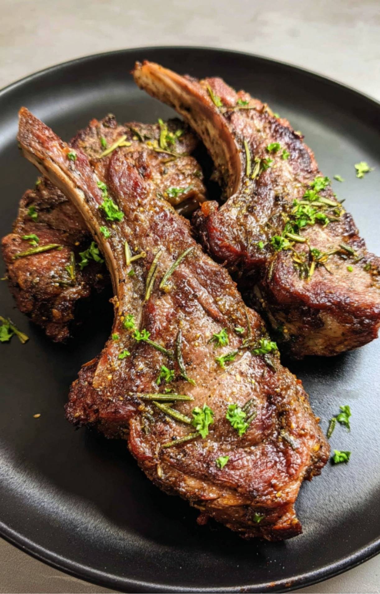 Three cooked air fryer lamb shoulder chops on a black plate, garnished with fresh rosemary and parsley.