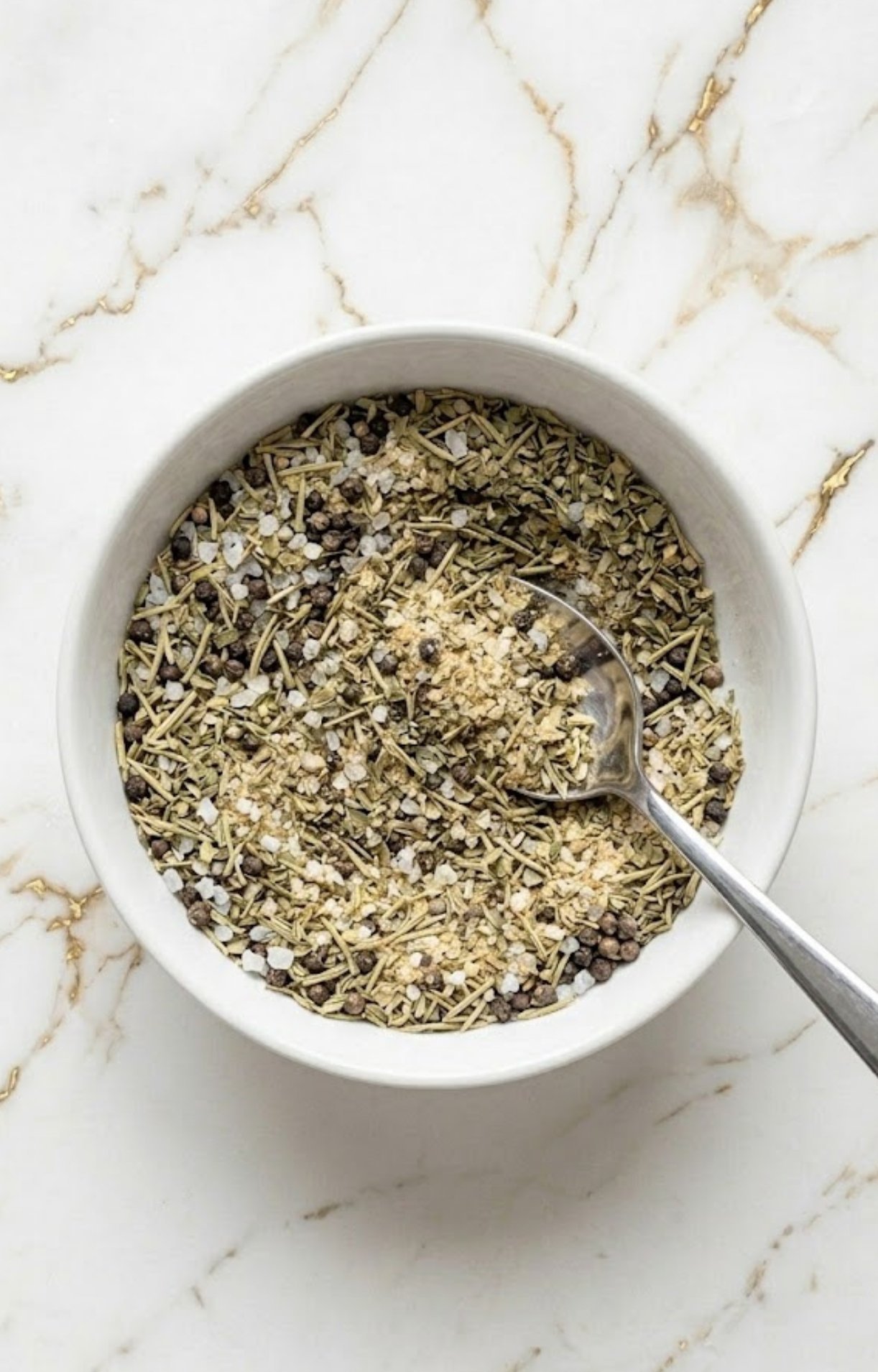 A silver spoon mixing together coarse salt, black peppercorns, and green herbs in a white bowl to create the Smoked Tomahawk Steak Rub.