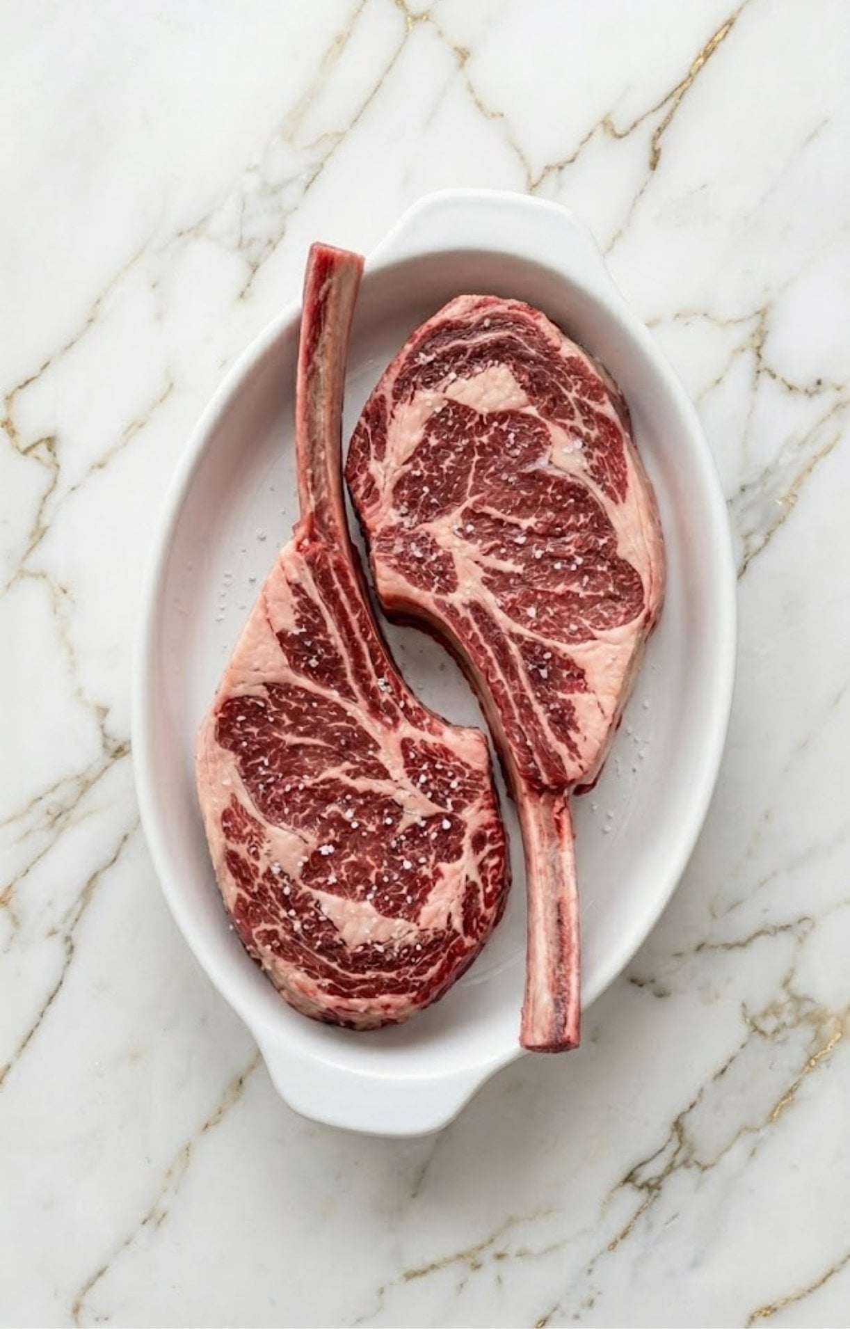 Two thick-cut raw tomahawk steaks resting in a white ceramic dish, lightly seasoned with large flakes of coarse sea salt to begin the dry brining process.
