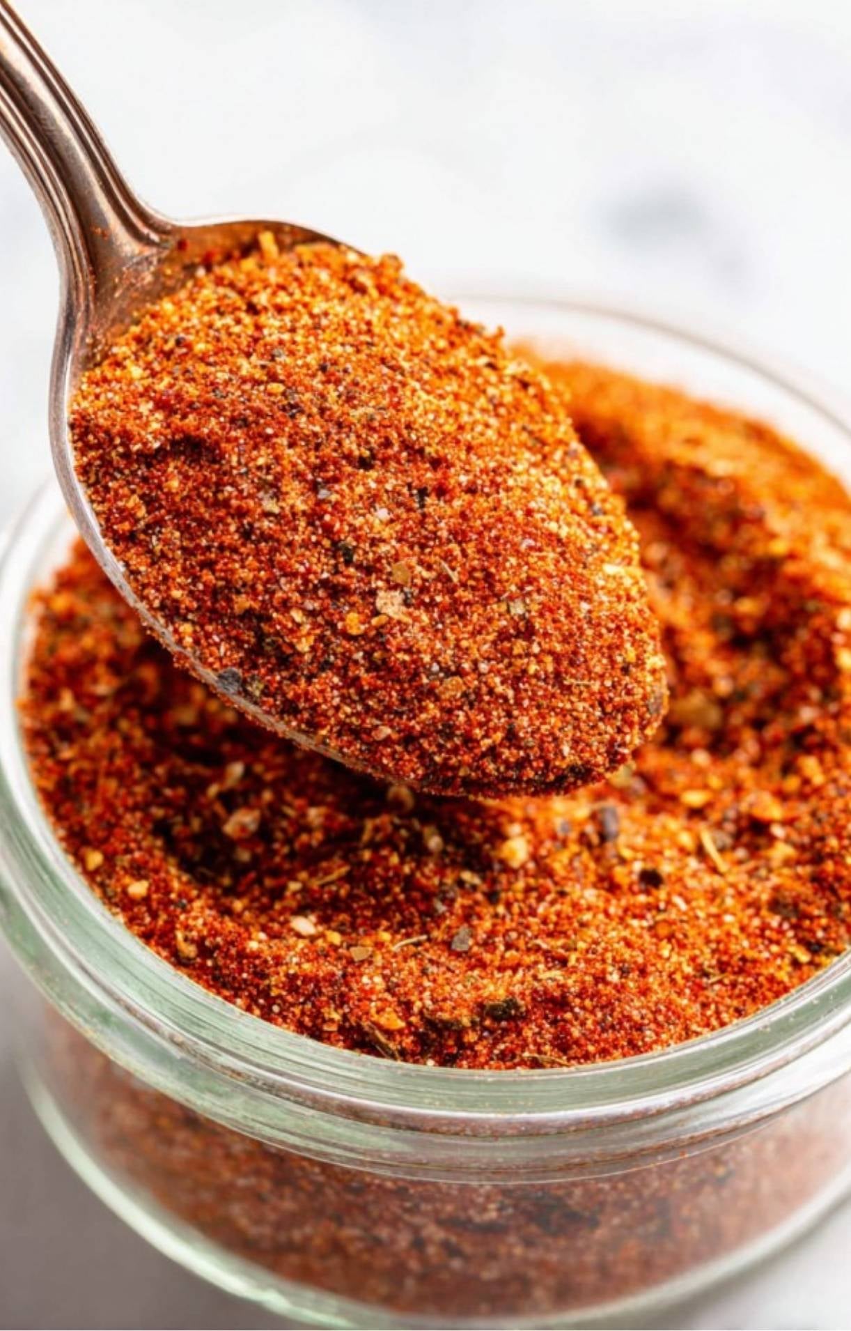 A silver spoon lifting a heap of textured smoked beef brisket rub from a glass jar, highlighting the coarse grains of pepper and spices.