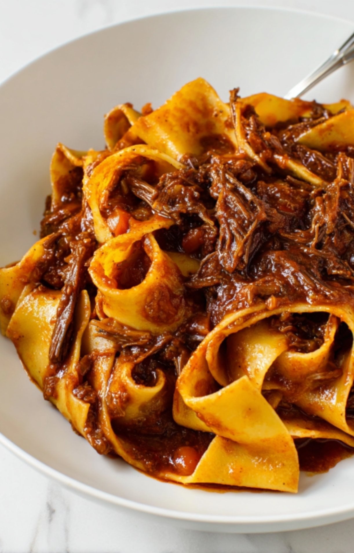 An extreme close-up of tender, shredded beef cheek ragu clinging to wide ribbons of pappardelle pasta in a white bowl.