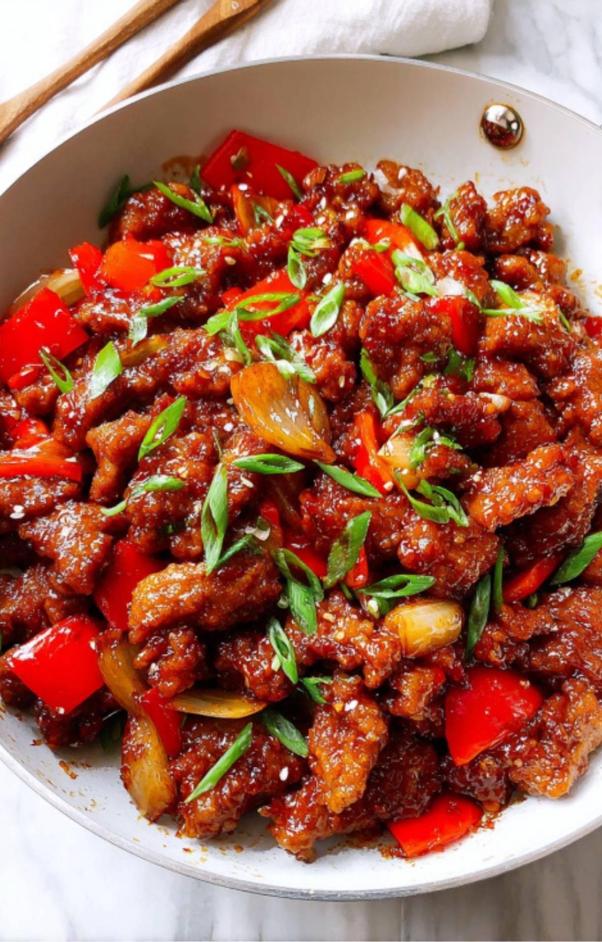 A large white skillet filled with glistening, sauce-coated crispy beef, sautéed onions, and vibrant red bell peppers garnished with scallions.
