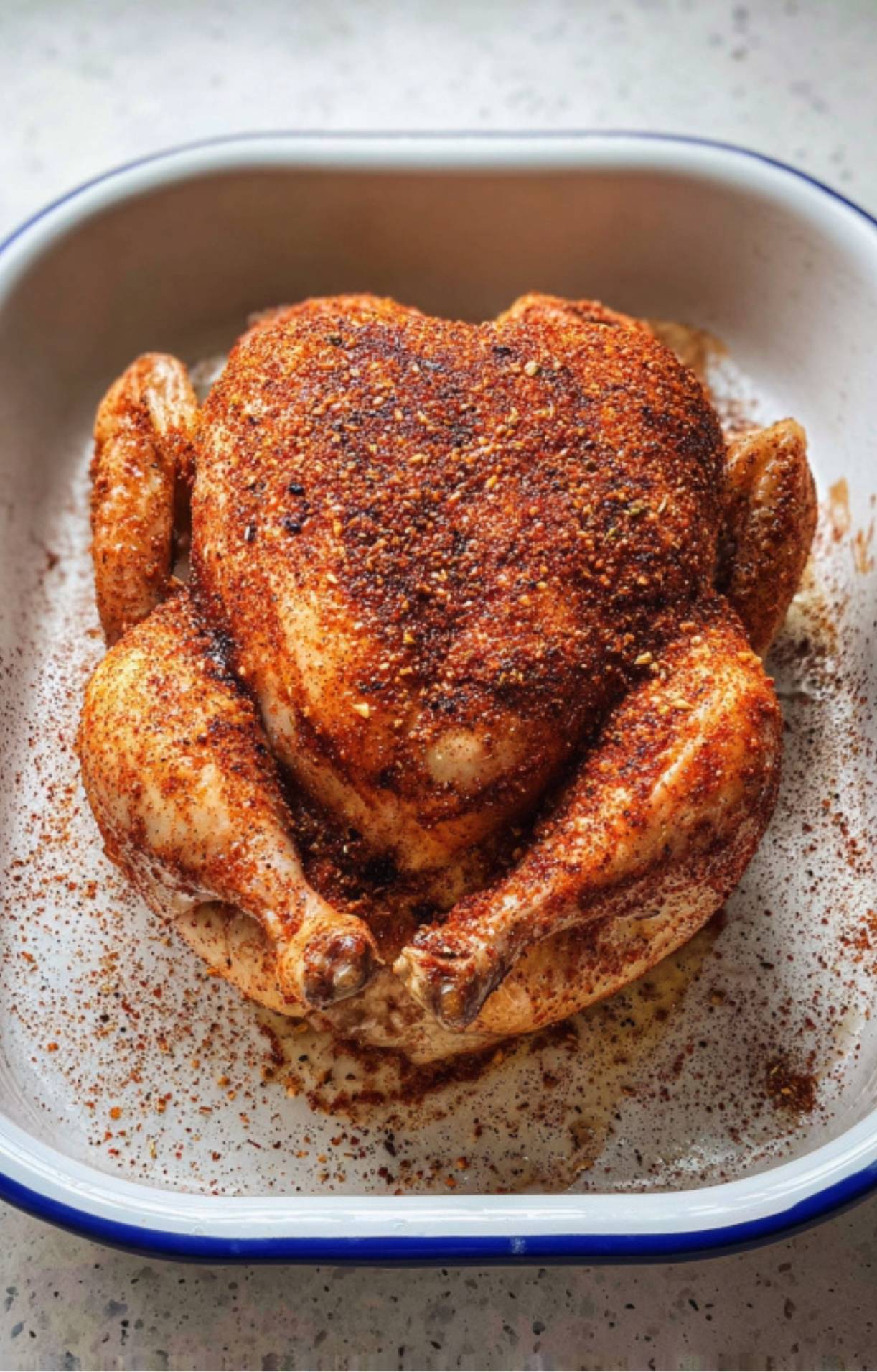 A whole raw chicken in a white roasting pan, generously coated in a red-toned dry spice rub.
