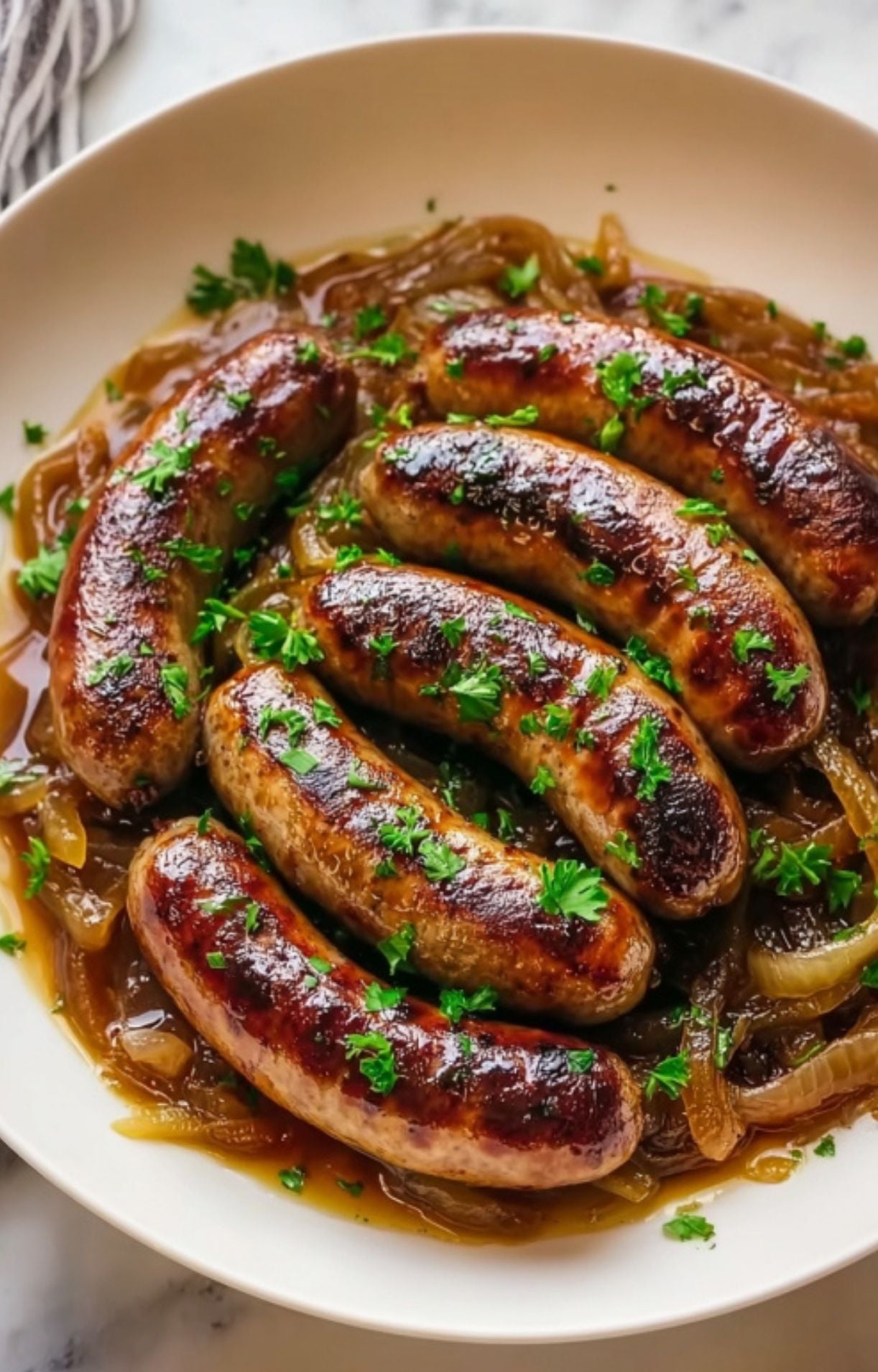 Six caramelized, golden-brown bratwurst sausages topped with fresh green parsley and served on a bed of savory sautéed onions in a shallow white bowl.