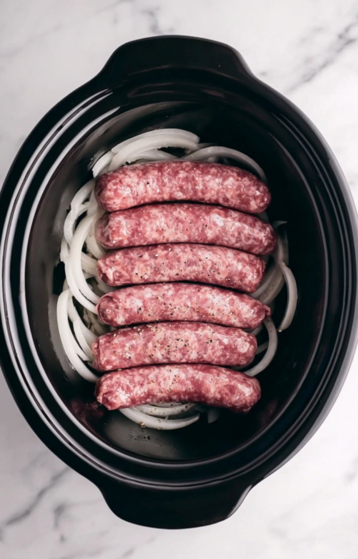 Raw bratwurst sausages neatly layered over a bed of sliced white onions in a black ceramic crockpot insert.