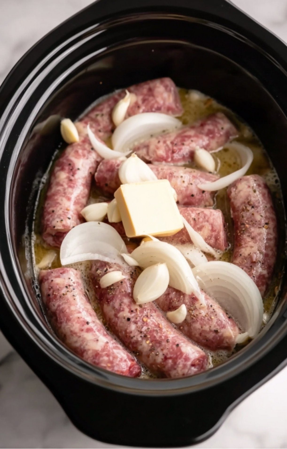 Raw sausages in a slow cooker topped with fresh garlic cloves, sliced onions, a pat of butter, and poured lager beer ready for cooking.