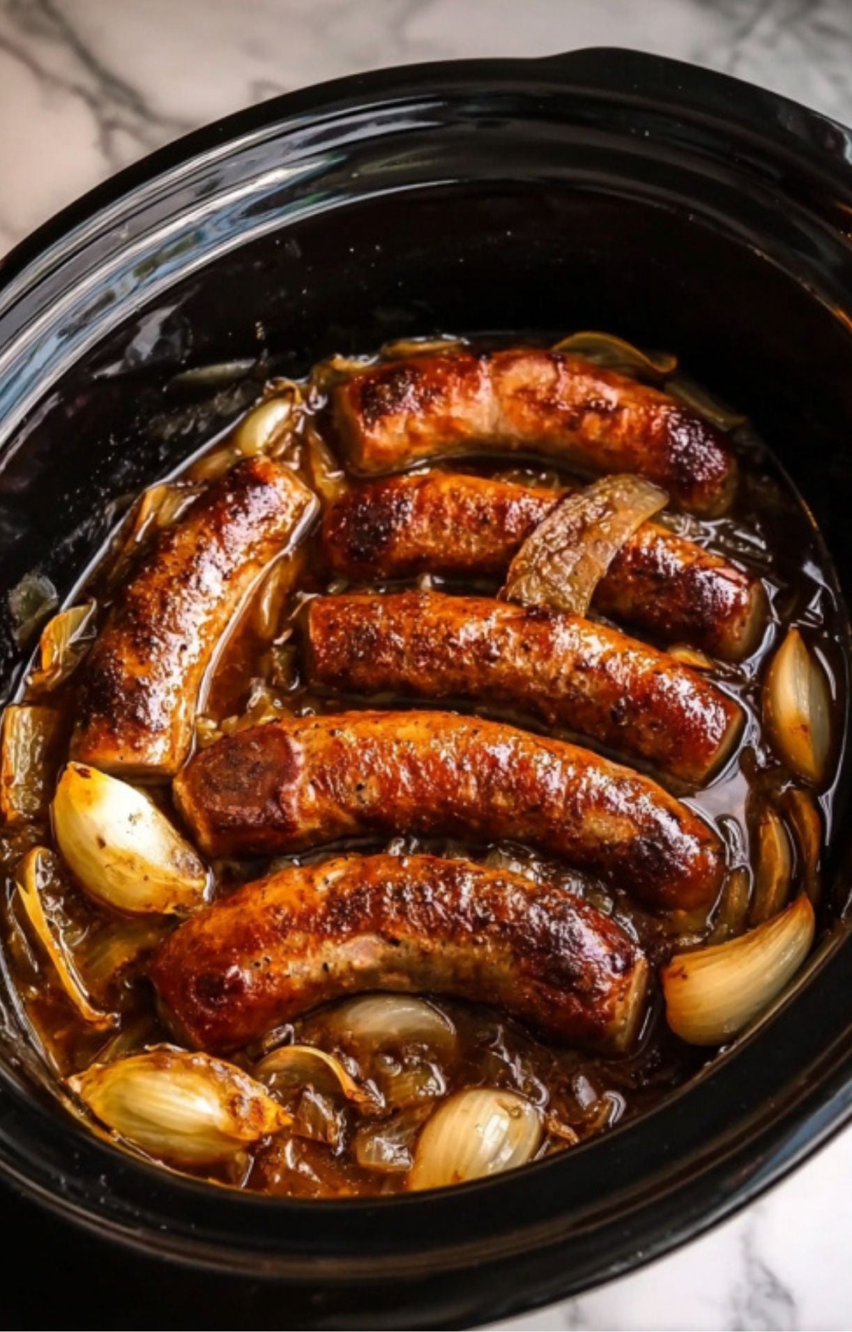 A top-down view of fully cooked, browned sausages resting in a savory beer and onion broth inside a black crockpot.