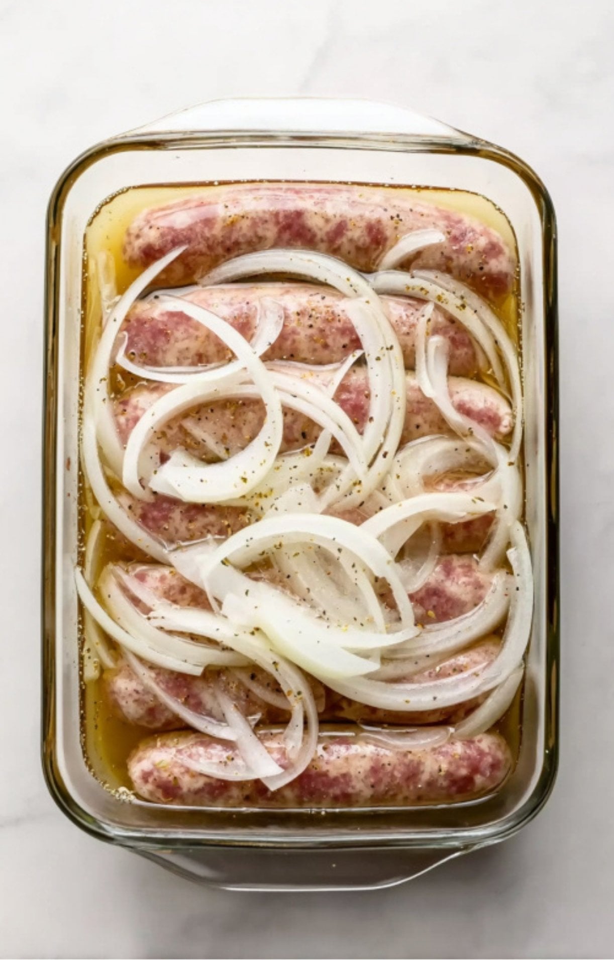 Raw bratwurst links and thick onion slices submerged in a beer marinade inside a glass rectangular baking dish.