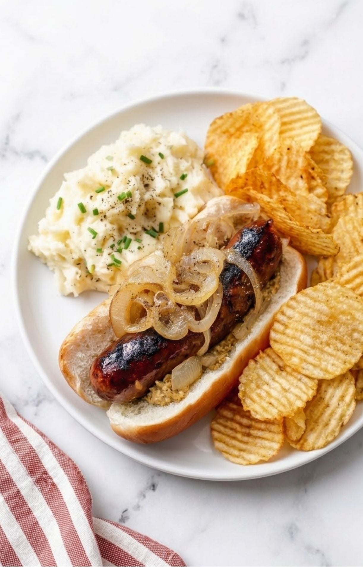 A loaded beer brat in a bun with mustard and onions, served with a side of creamy mashed potatoes and ruffled potato chips.