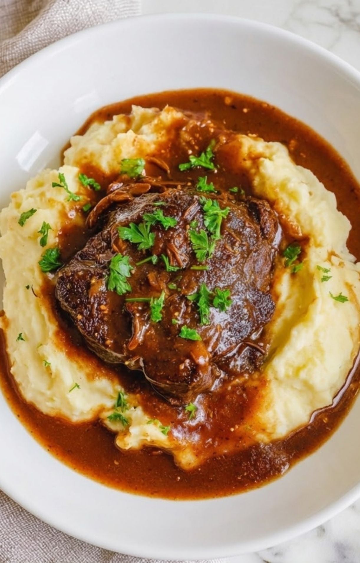 A top-down view of a tender braised beef cheek nestled in a well of creamy mashed potatoes, drizzled with rich red wine gravy and topped with fresh parsley.