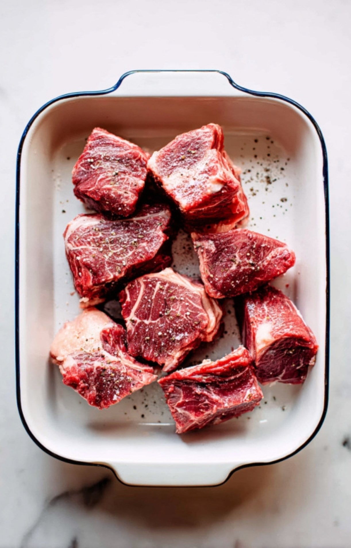 Large chunks of raw, bright red beef cheek seasoned with salt and black pepper, arranged in a white rectangular baking dish on a marble surface.