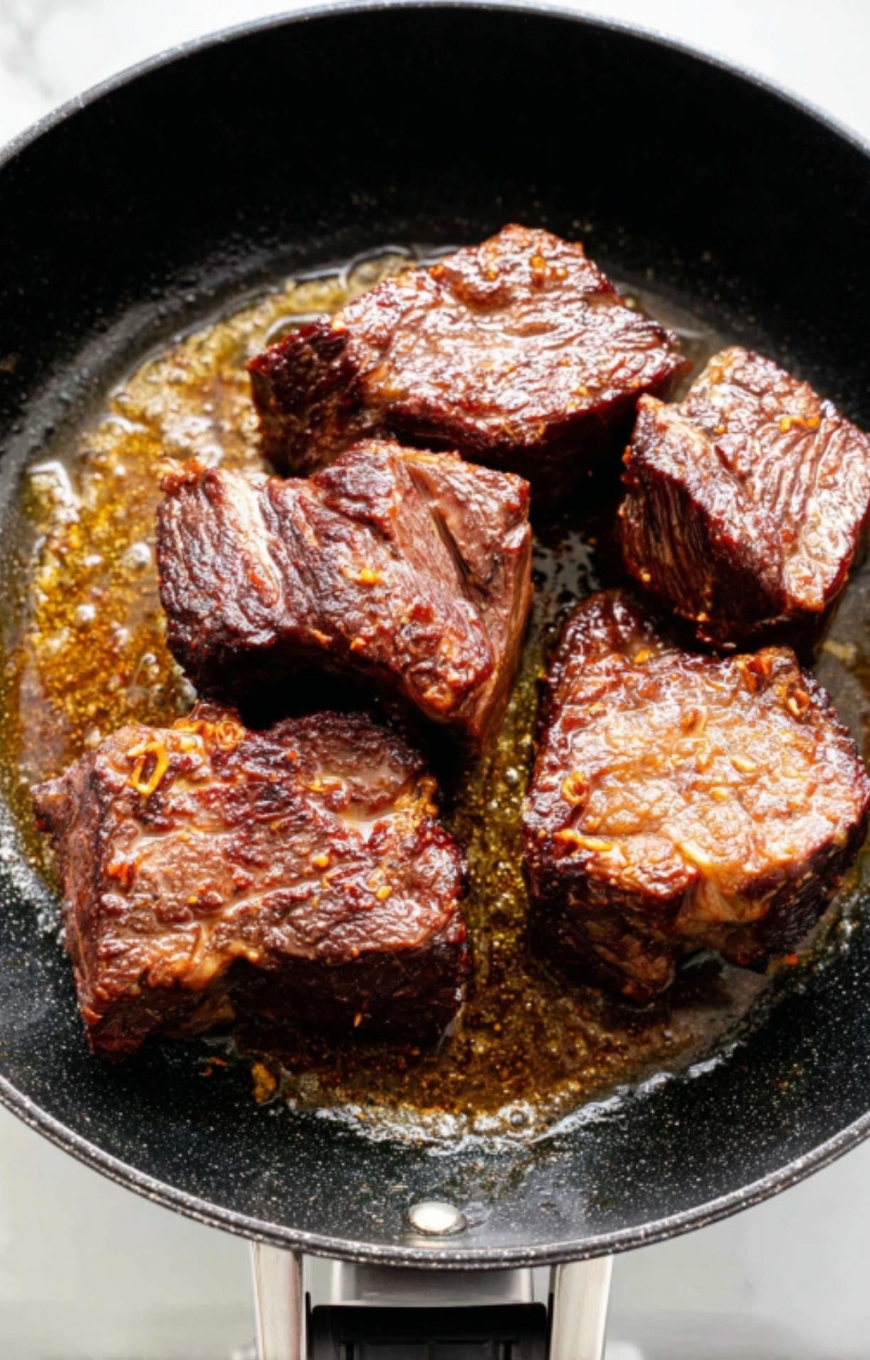 Thick chunks of seasoned beef cheek being seared until deep brown in a black skillet with shimmering oil.