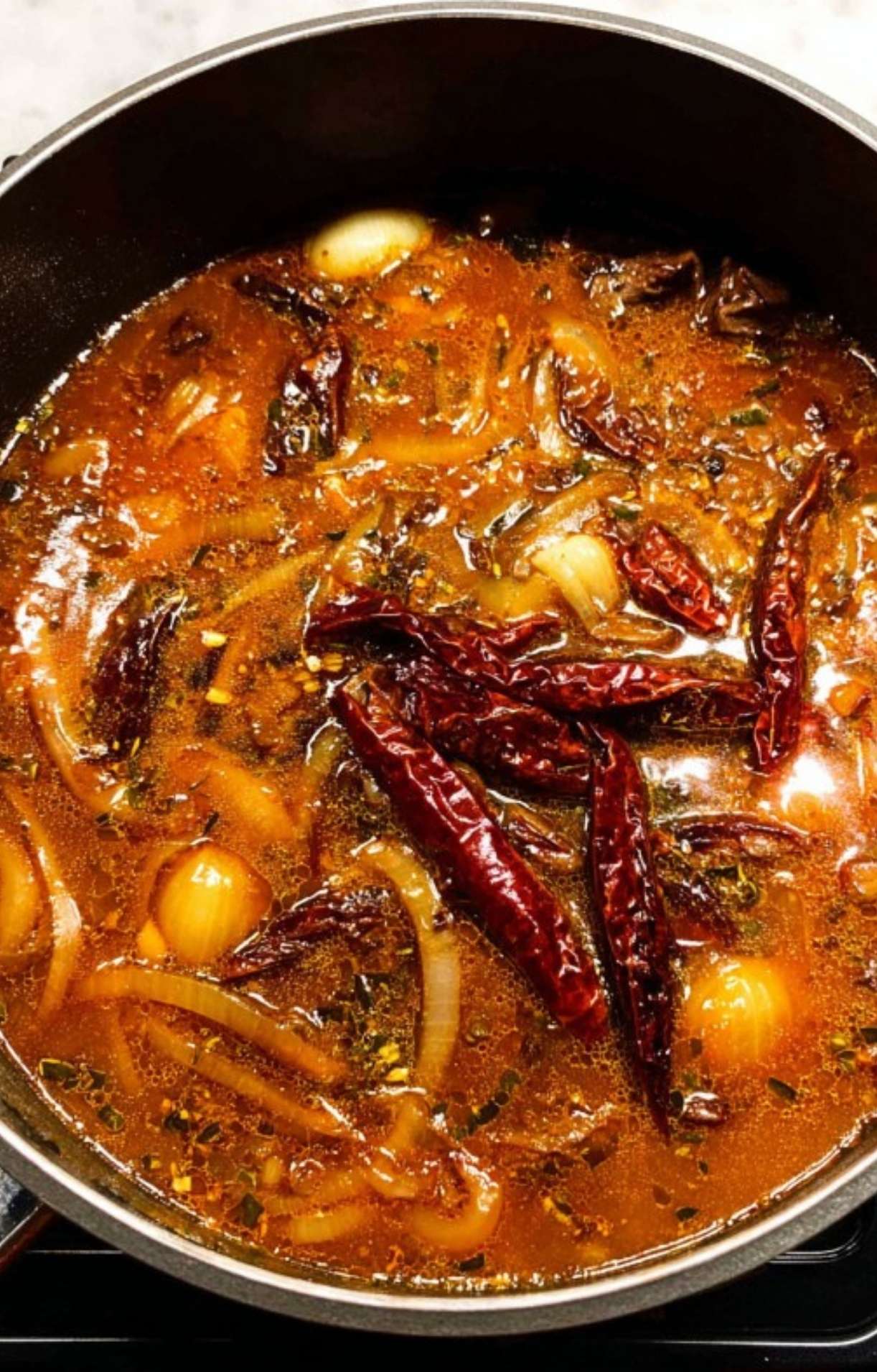 Whole dried chiles, onions, and garlic simmering in a rich, reddish-brown broth within a large pot.