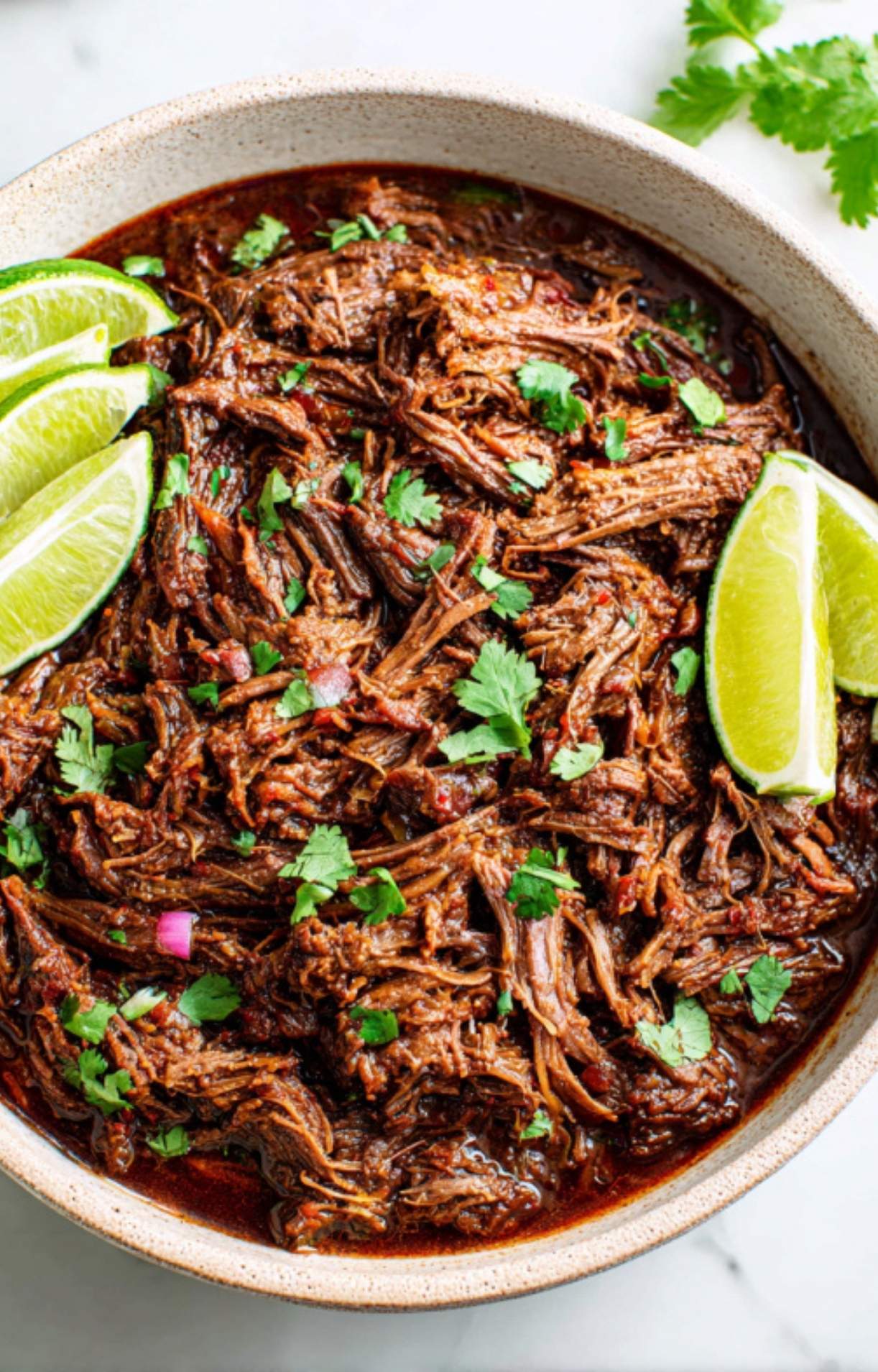A large ceramic bowl filled with juicy shredded beef cheek, garnished with fresh cilantro, diced red onions, and lime wedges.