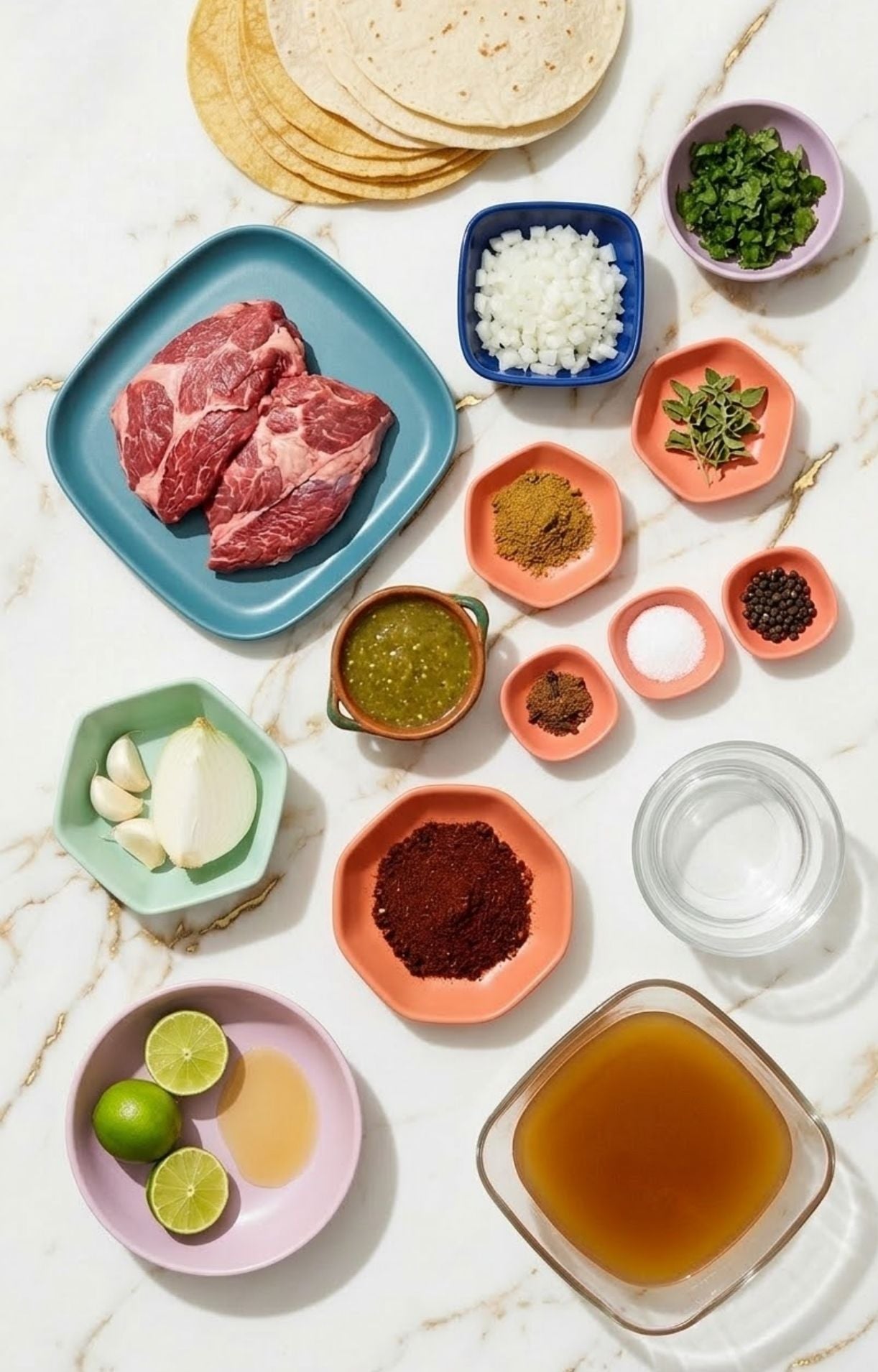 An overhead view of raw beef cheeks, tortillas, spices, onions, limes, and broth arranged on a marble surface.