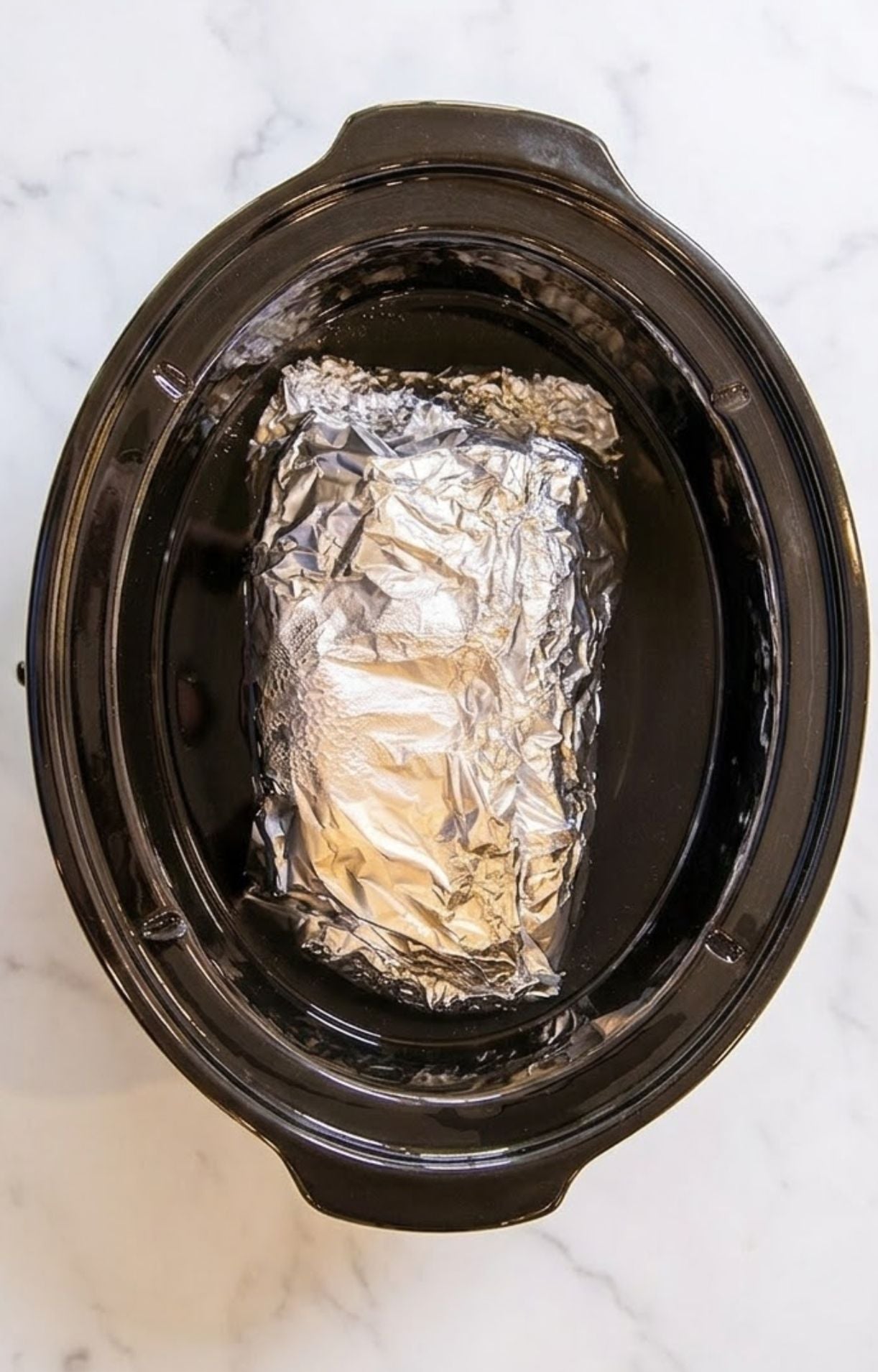 A tight foil packet containing the seasoned beef placed inside a black slow cooker insert.