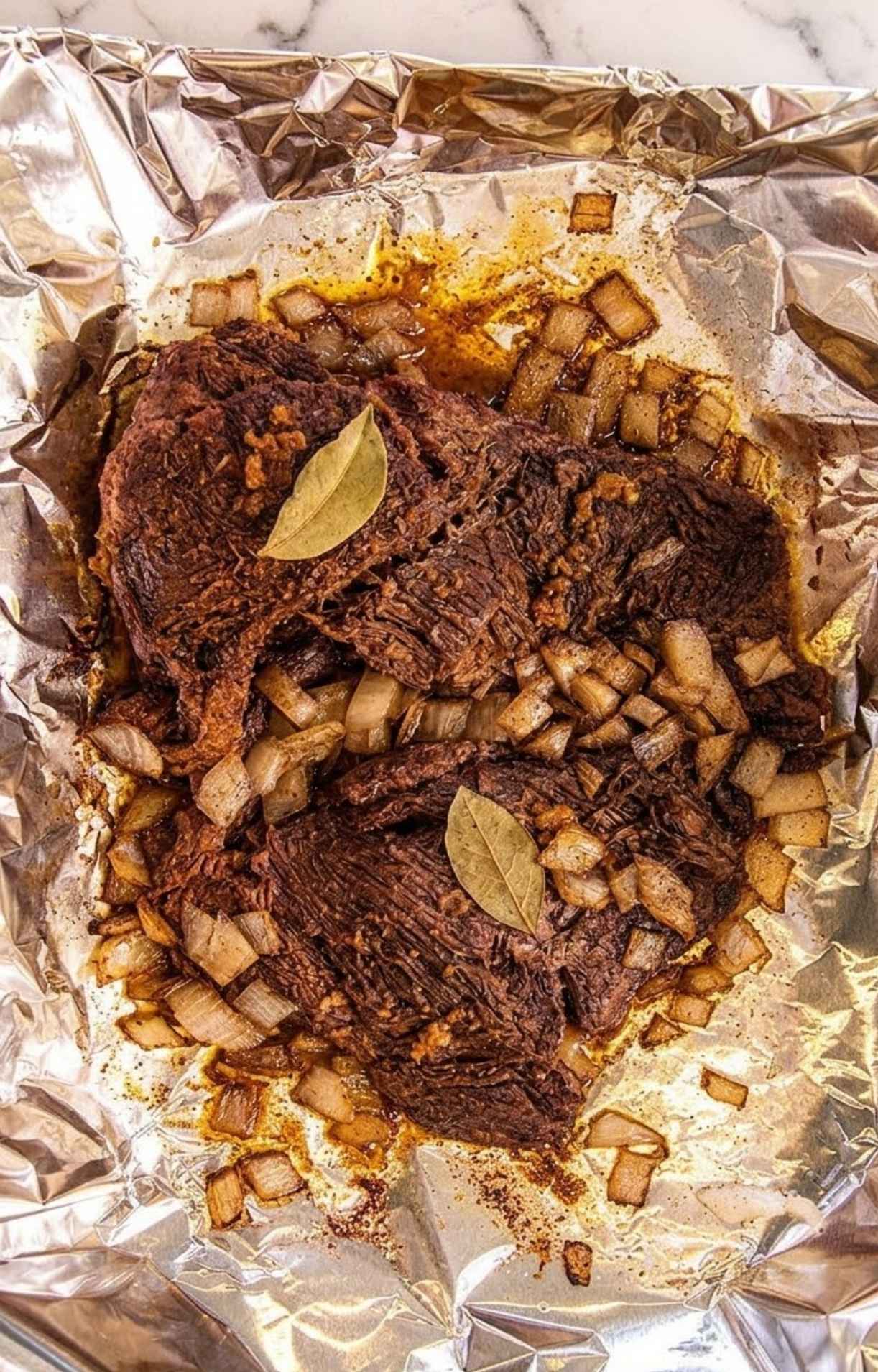 Tender, cooked beef cheeks resting on foil surrounded by softened, translucent diced onions and bay leaves.