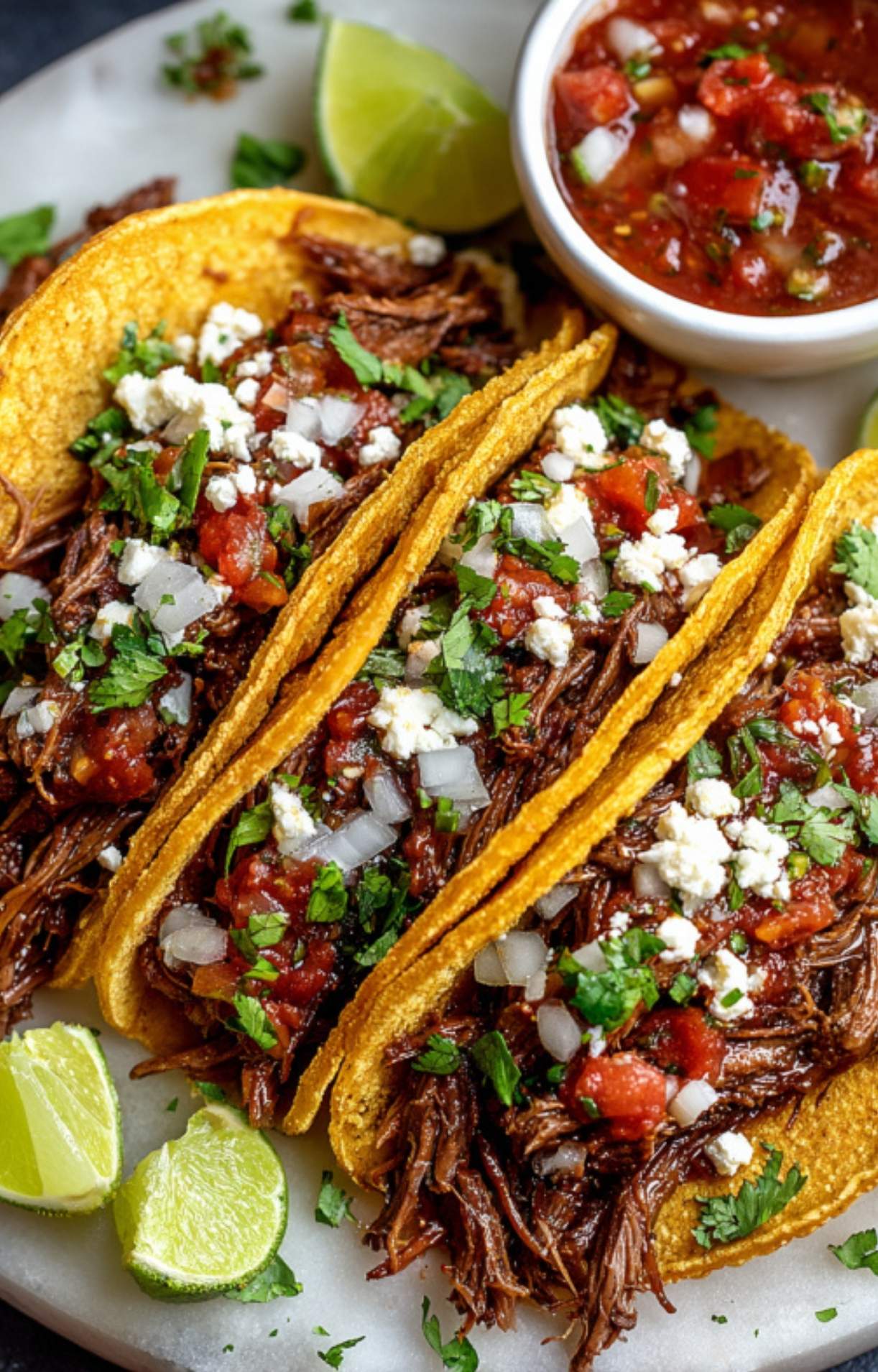 Three corn tortillas filled with shredded beef, topped with salsa, white cheese, onions, and cilantro.