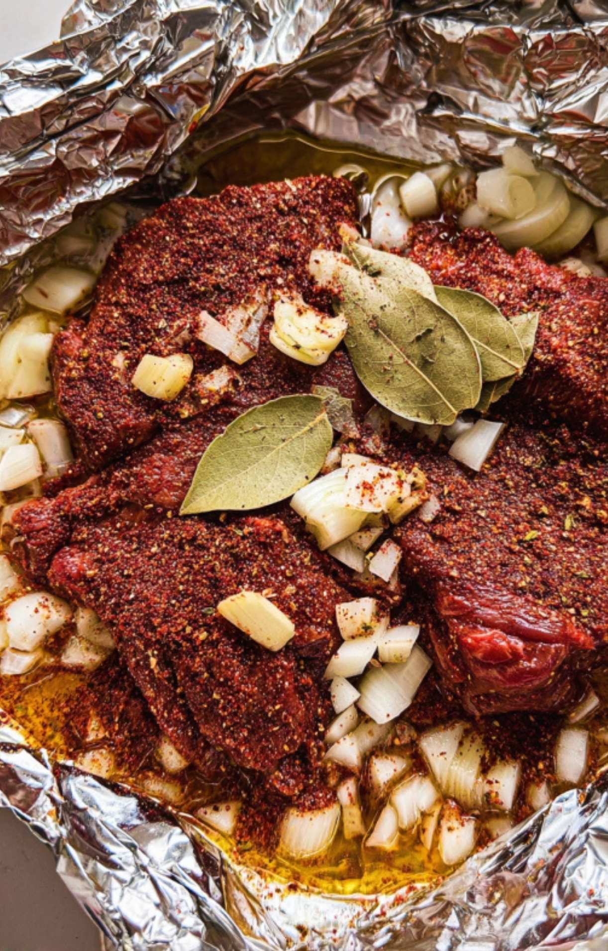A close-up of raw beef cheeks heavily coated in a red spice rub, layered with diced white onions, fresh bay leaves, and garlic cloves inside a foil-lined roasting pan.