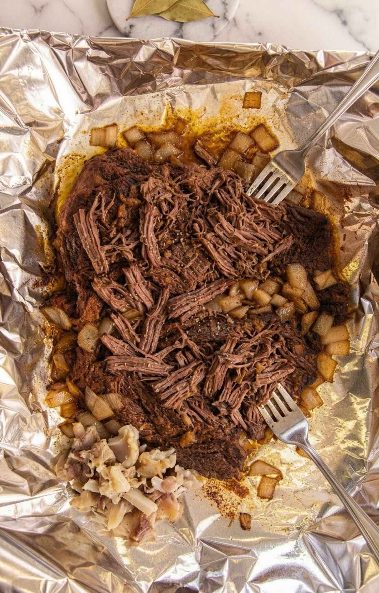 Two silver forks pulling apart a tender, slow-cooked beef cheek on a sheet of crumpled aluminum foil, surrounded by caramelized onions and rendered fat.