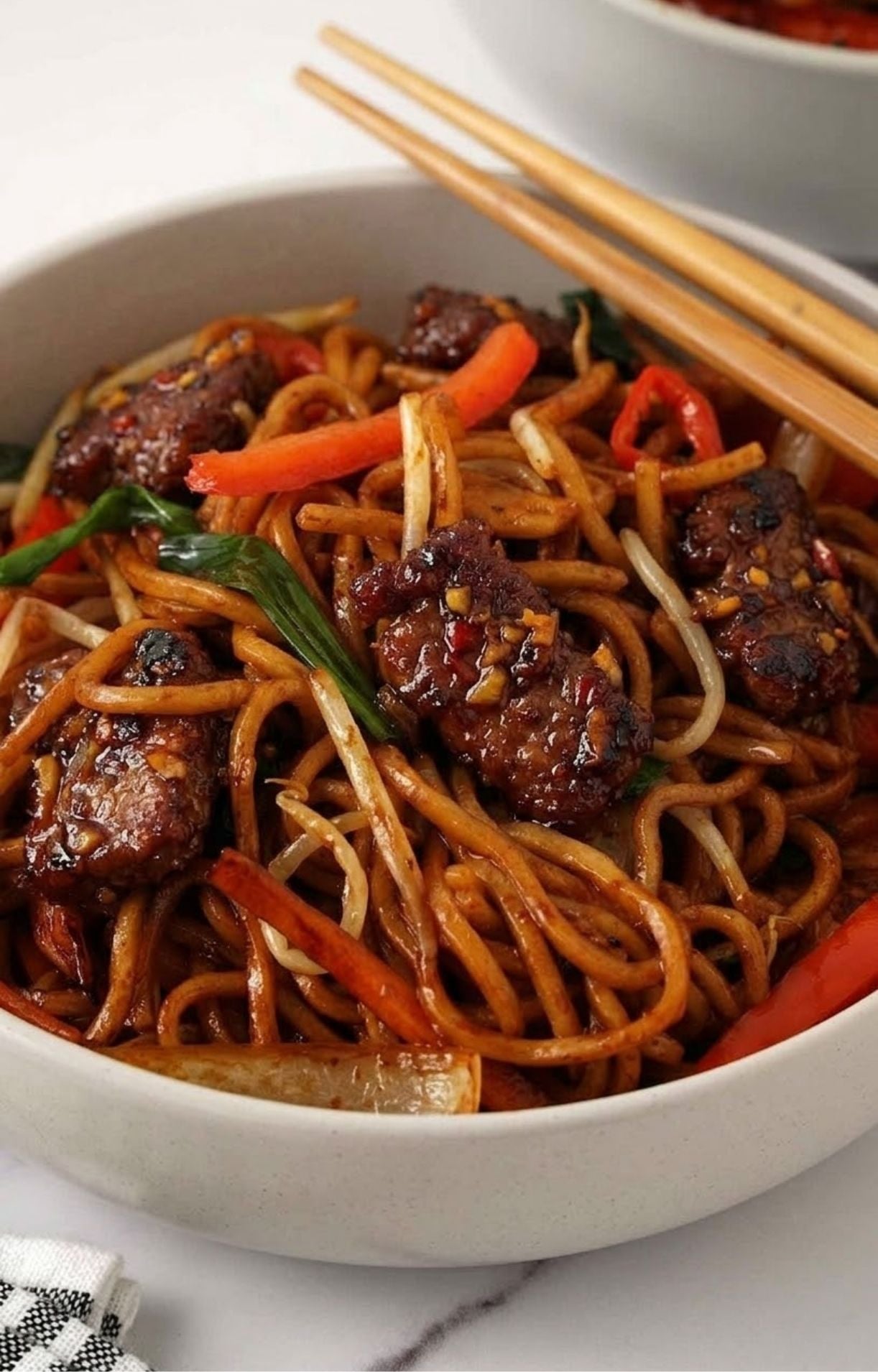 A close-up shot of a bowl of glossy lo mein noodles tossed with crispy glazed beef, red peppers, and green onions, with wooden chopsticks resting on the side.