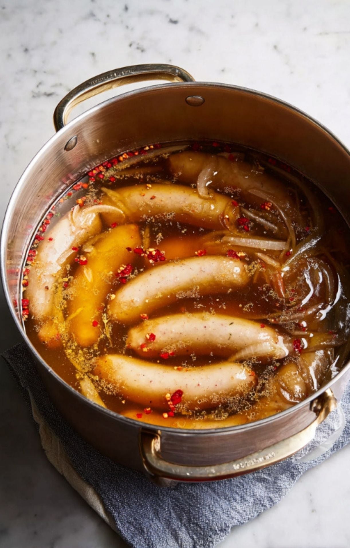 Raw bratwurst sausages and sliced onions simmering in a flavorful beer and red pepper flake liquid in a large metal pot.