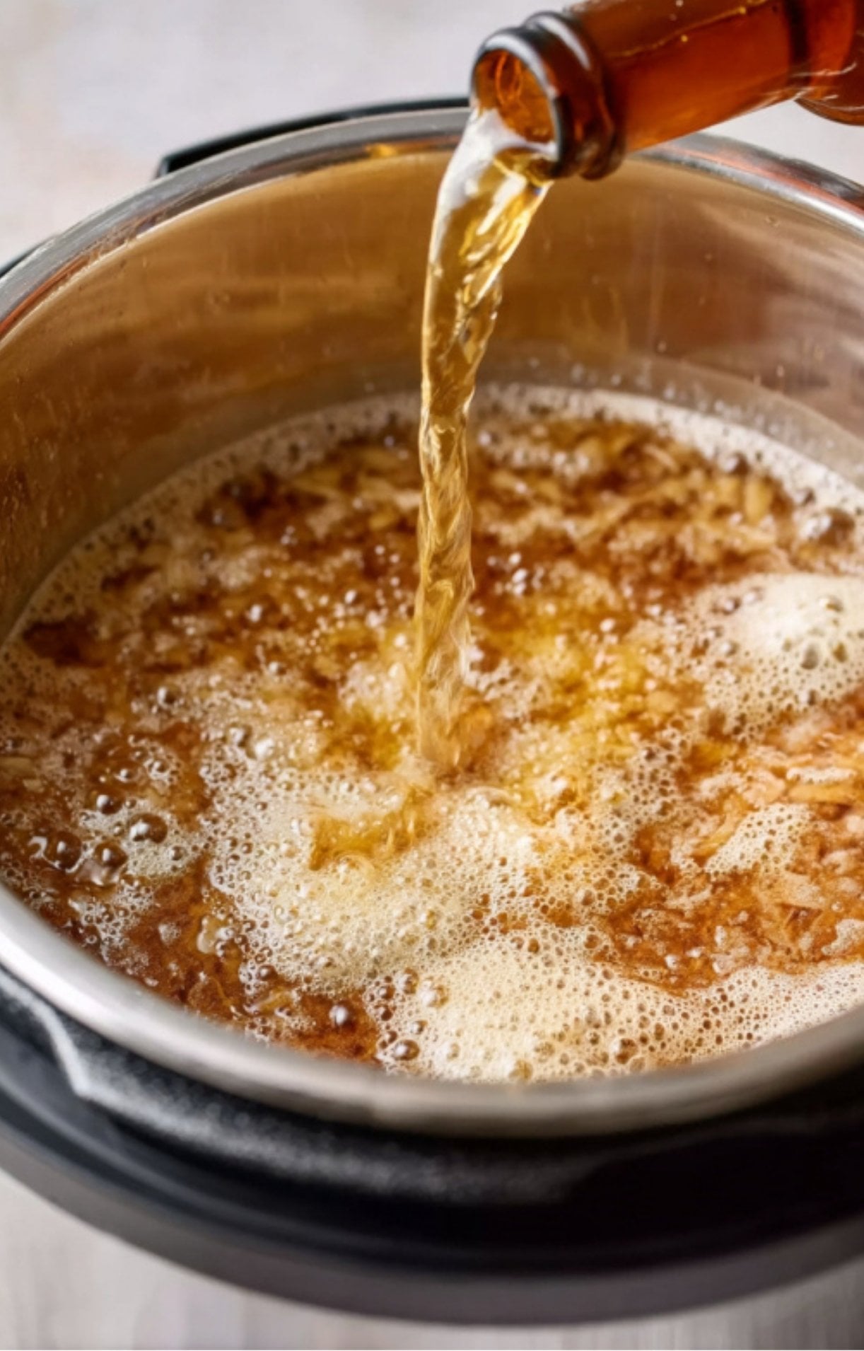 Light beer being poured from a bottle into a rice cooker pot filled with sautéed onions and bratwurst.