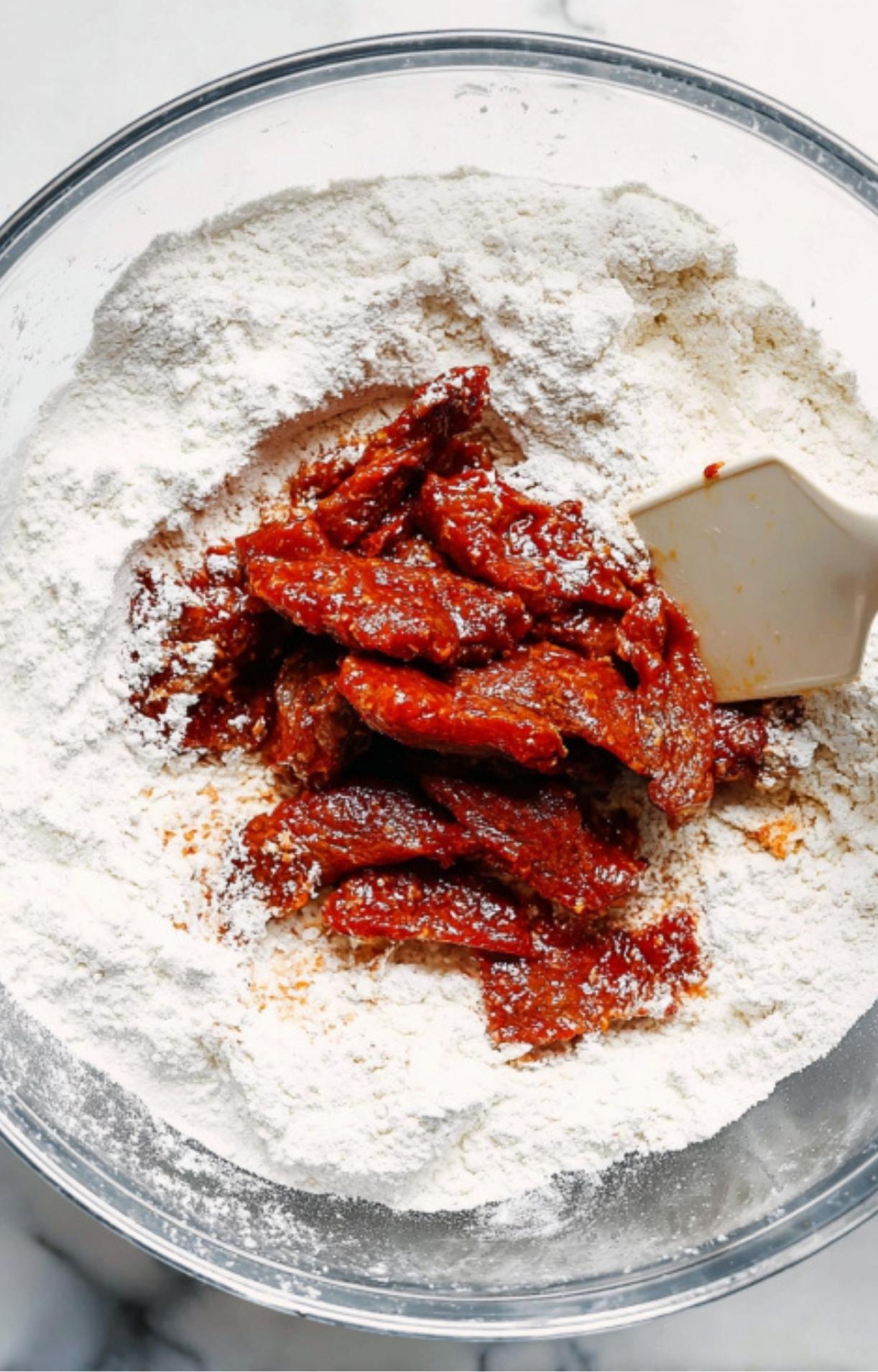 Marinated beef strips being tossed in a bowl of white flour and cornstarch mixture with a spatula to create a crispy coating.