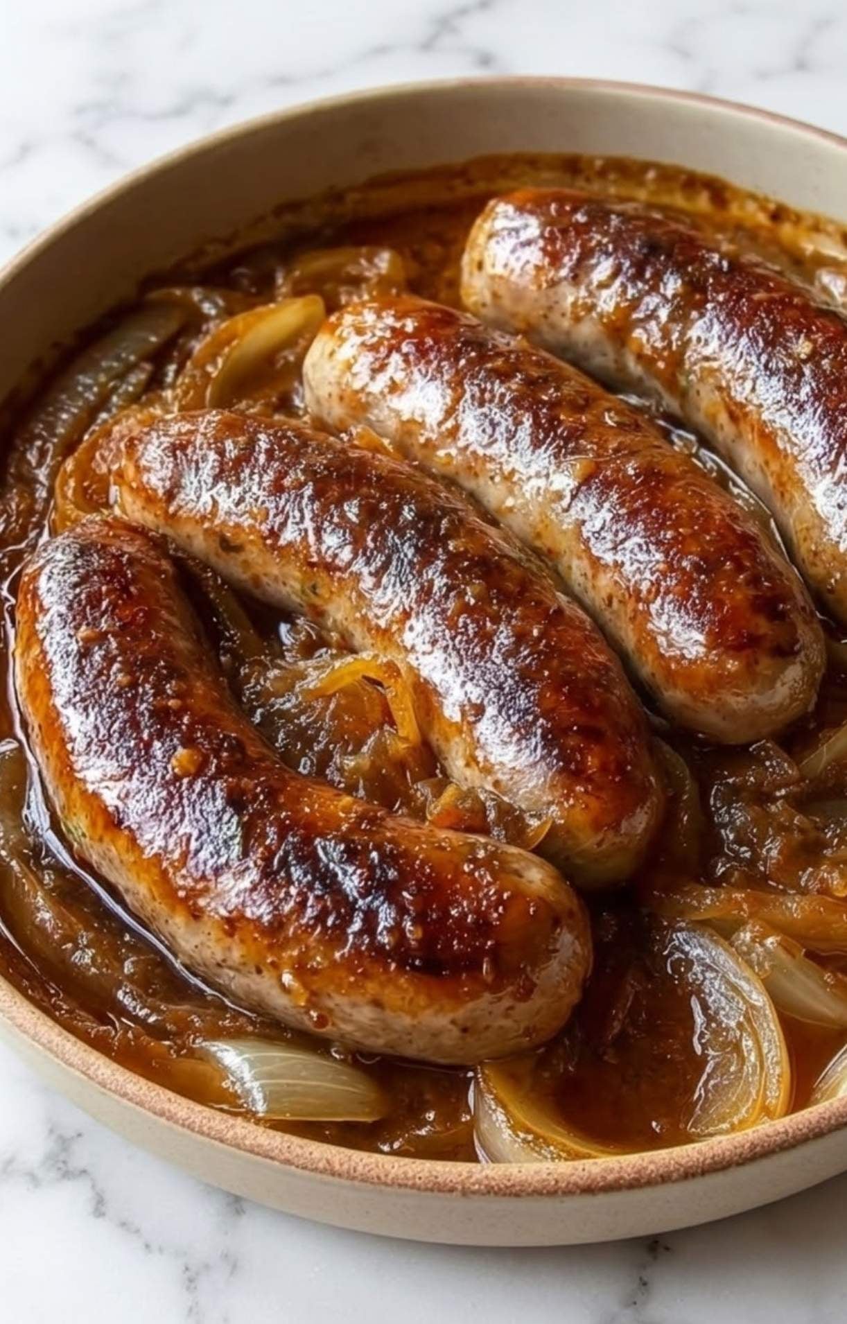 Three juicy, browned bratwurst sausages resting on a thick bed of jammy, caramelized onions in a shallow serving dish.