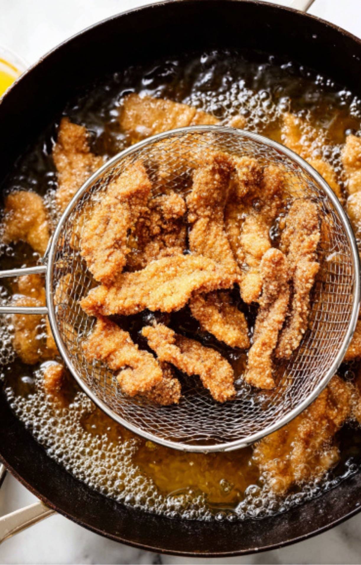 A metal spider strainer lifting golden-brown, deep-fried beef strips out of a pot of bubbling hot oil.