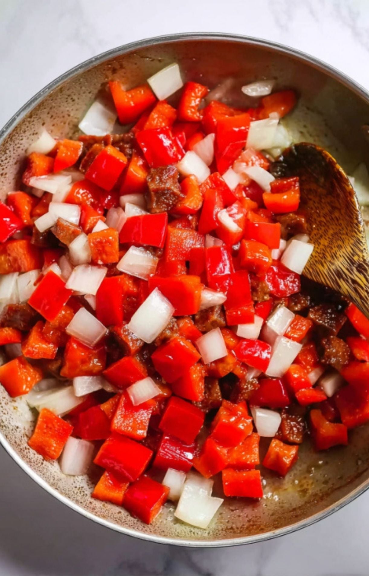 Diced red bell peppers and white onions being tossed in a pan with a wooden spoon until slightly softened.