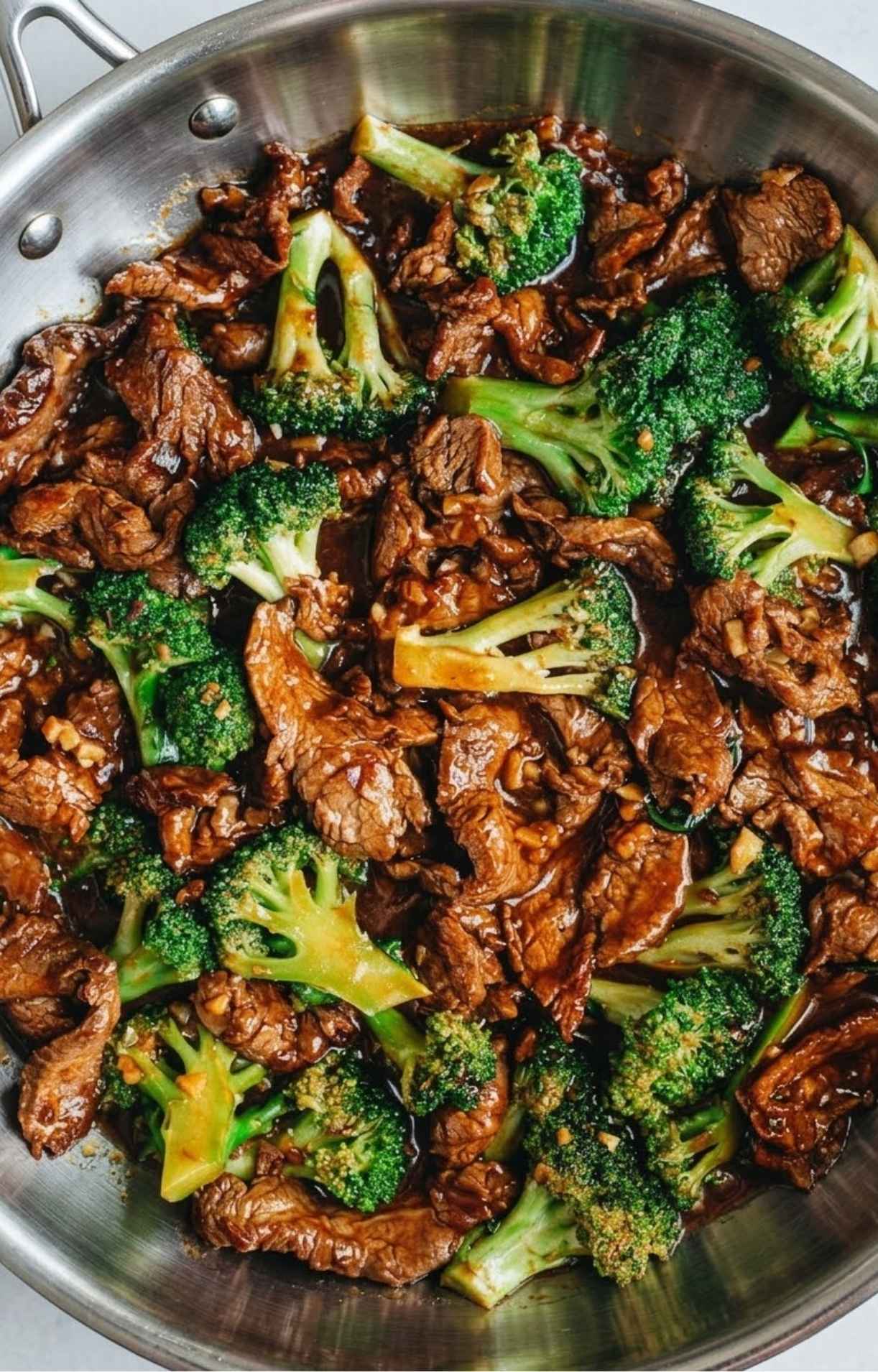 Beijing beef and broccoli simmering in a stainless steel skillet, highlighting the rich, dark stir-fry sauce.