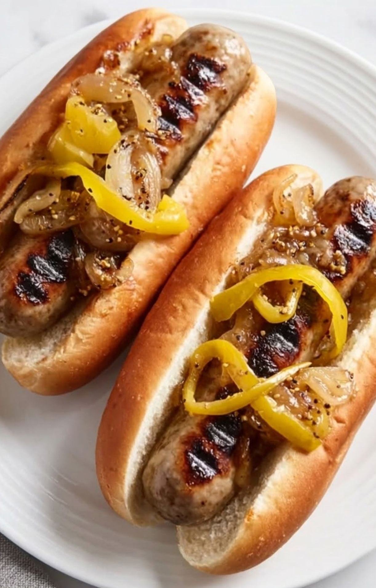 Macro shot of two beer-boiled brats with perfect grill marks, loaded with onions and bright yellow peppers.