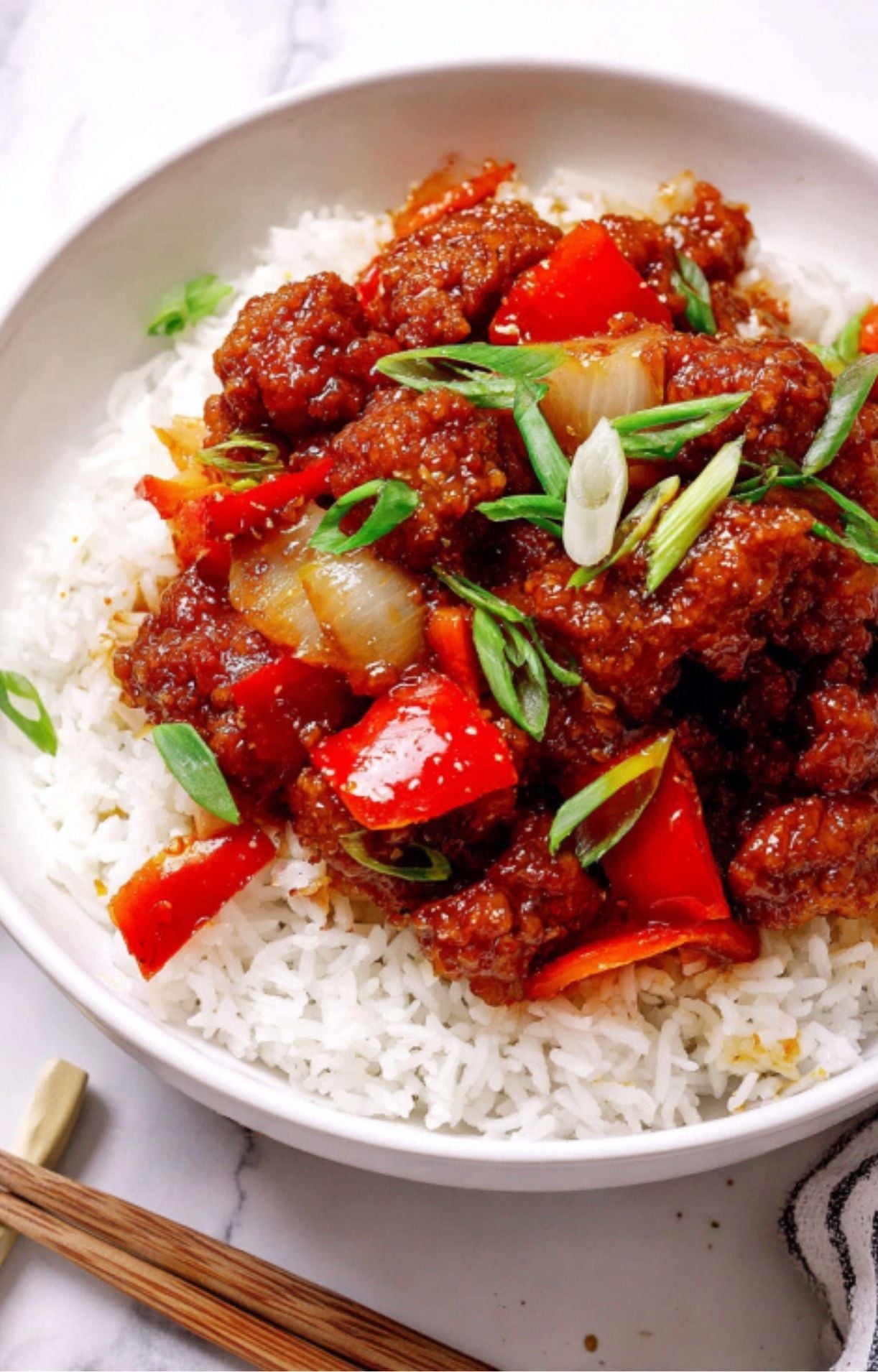 A white bowl filled with a generous serving of steamed white rice topped with crispy, saucy Beijing beef, large chunks of red bell pepper, and fresh sliced green onions.