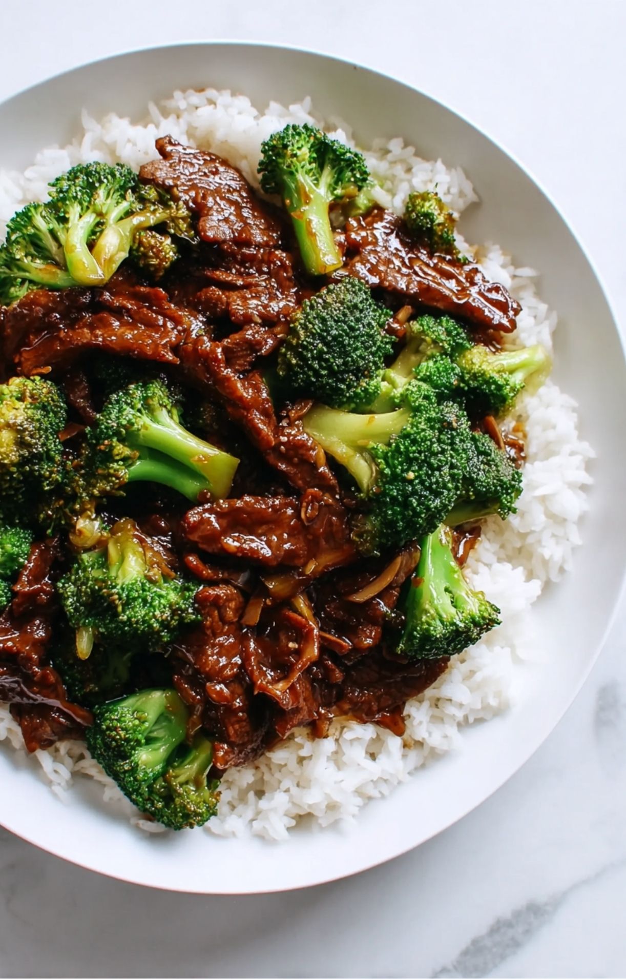 A top-down view of a generous portion of Beijing beef and broccoli served over steamed rice in a white bowl.
